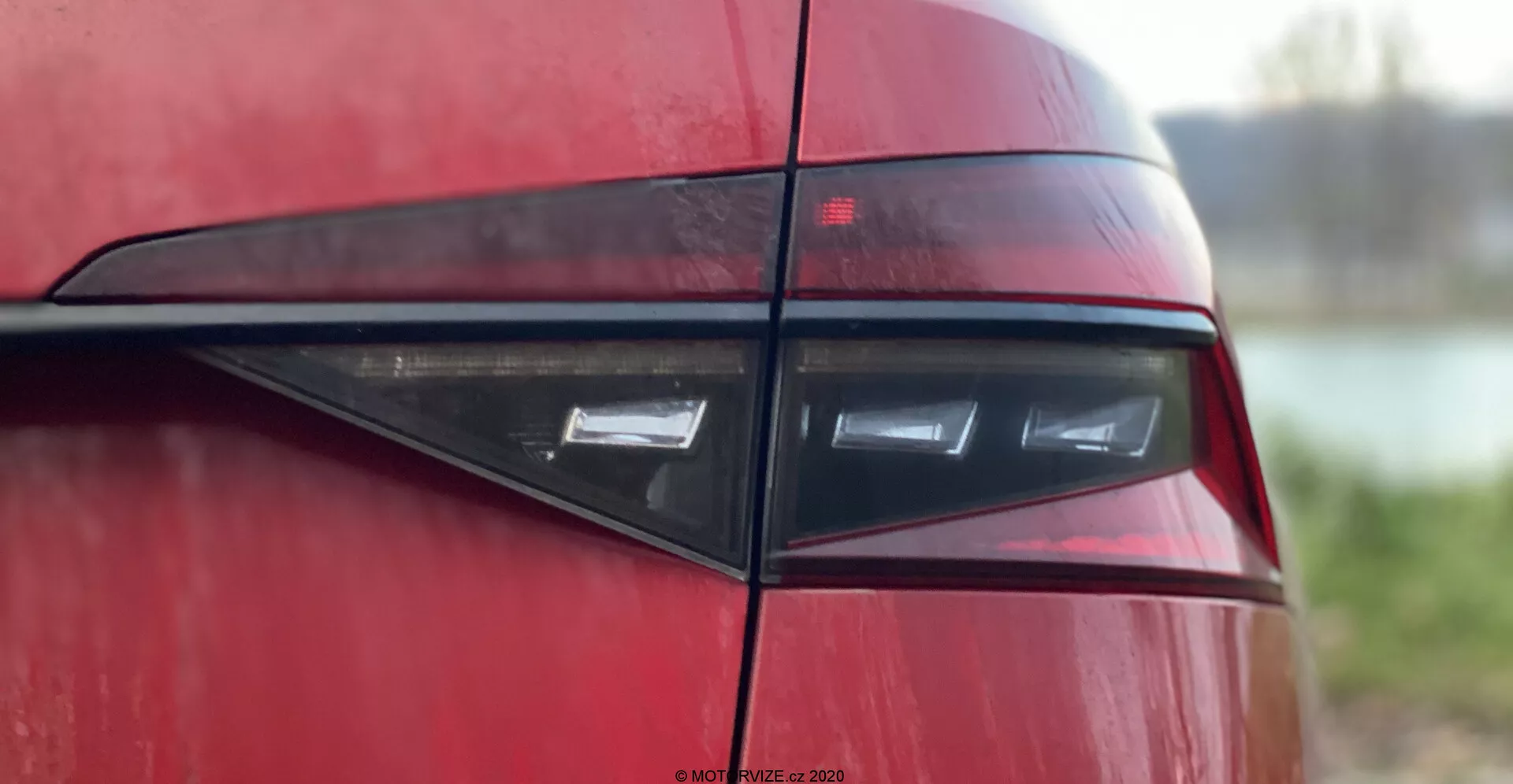 Detailný záber na zadnú časť vozidla Skoda Superb 2019, zameraný na zadné svetlo z bočného uhla, ktorý zobrazuje moderný, elegantný dizajn s horizontálnymi LED prvkami v tmavočervenom kryte, odrážajúci súčasnú športovú alebo luxusnú estetiku vozidiel. Lesklá karoséria a čierna horizontálna lišta zvyšujú vizuálnu príťažlivosť na rozmazanom pozadí.