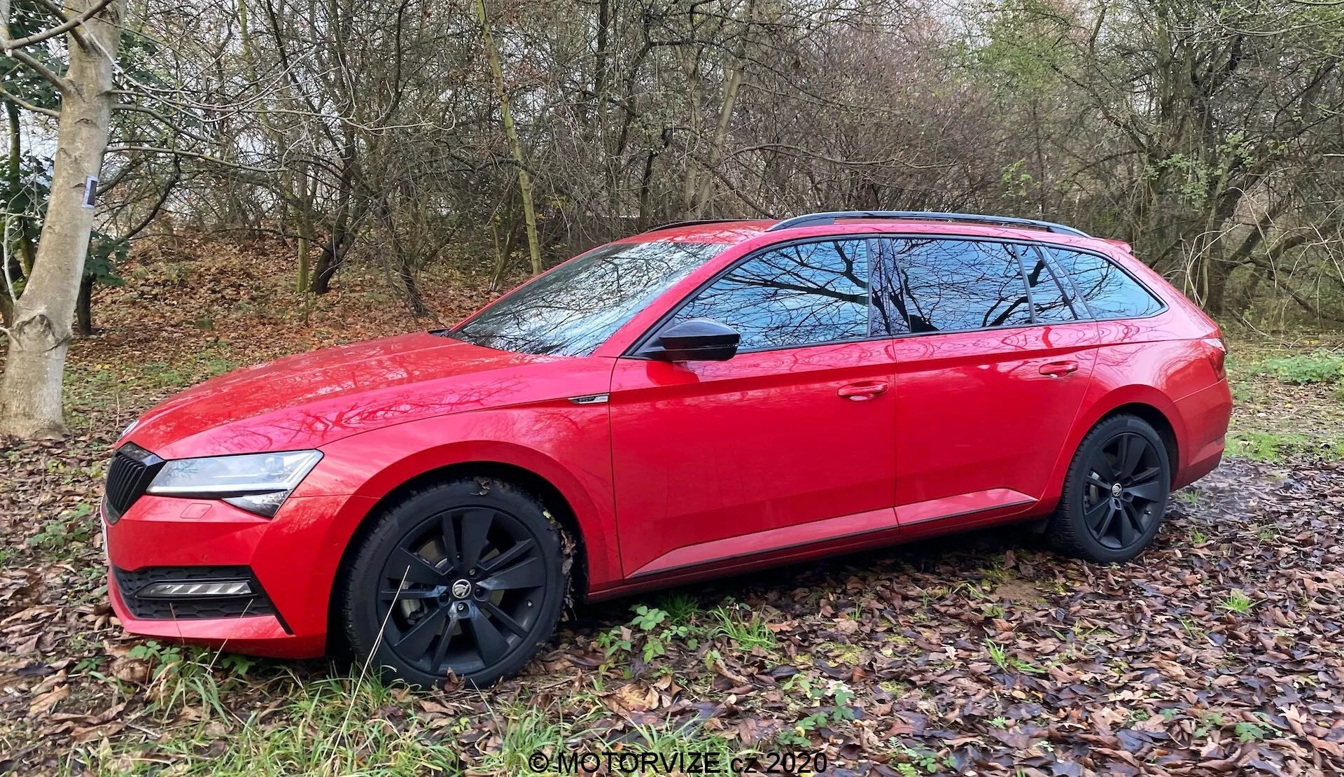 Bočný profil červeného vozidla Škoda Superb Limousine 2019 zaparkovaného na trávnatej ploche s listami na zemi. Dizajnové prvky vozidla zahŕňajú čierne zliatinové kolesá, okná s chrómovým rámom, spätné zrkadlá vo farbe karosérie a strešné lišty. Obrázok zachytáva dĺžku vozidla, ukazuje jeho kombi tvar, elegantný moderný dizajn a kontrast medzi červenou karosériou a čiernymi kolesami.