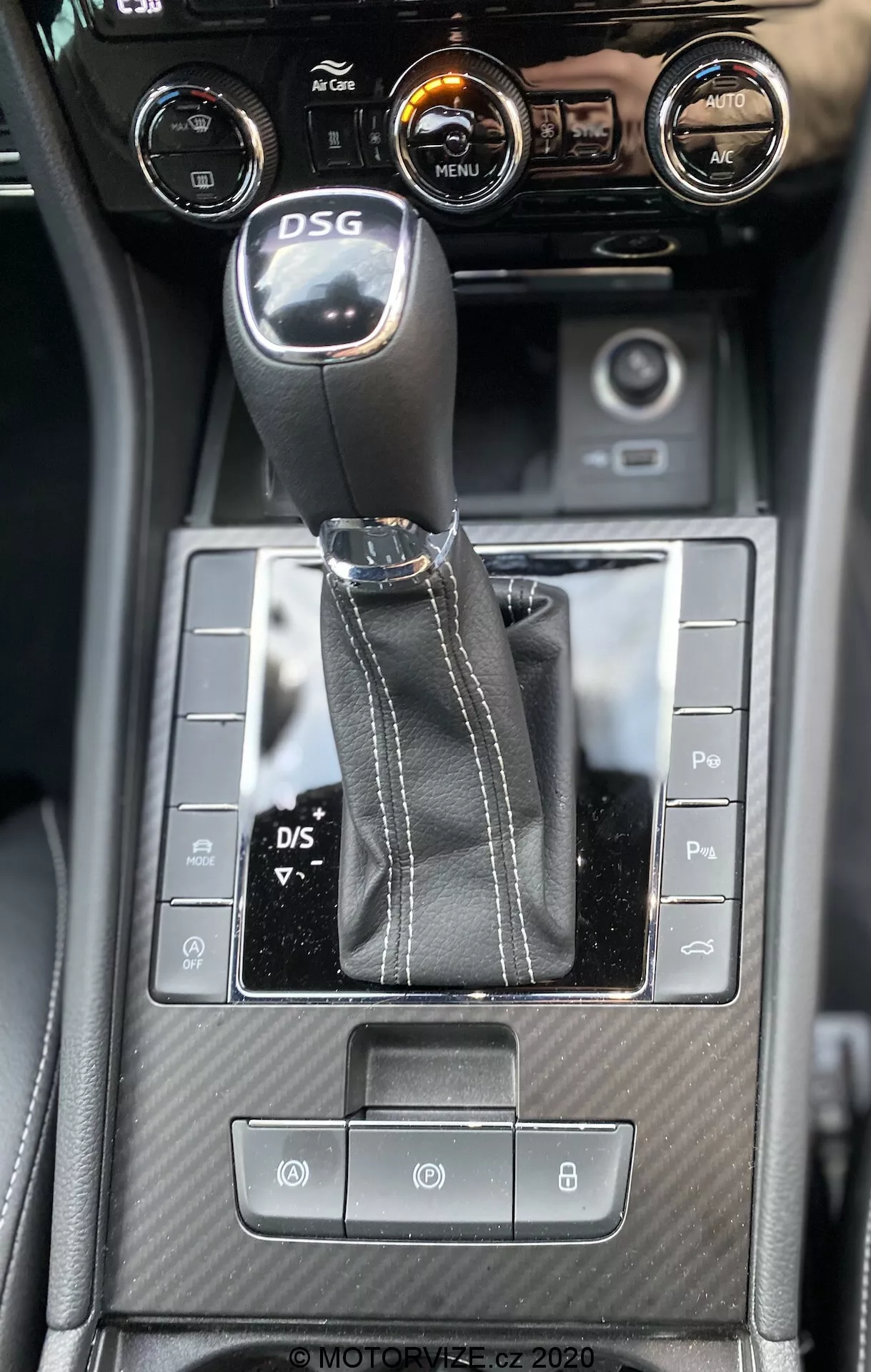Pohľad na interiér stredovej konzoly Škoda Superb 2019 zhora, zvýrazňujúci DSG radiacu páku, symetrické ovládacie tlačidlá, textúrované detaily pripomínajúce karbón a ovládanie klimatizácie s centrálnym dotykovým displejom zobrazujúcim informácie o vozidle.