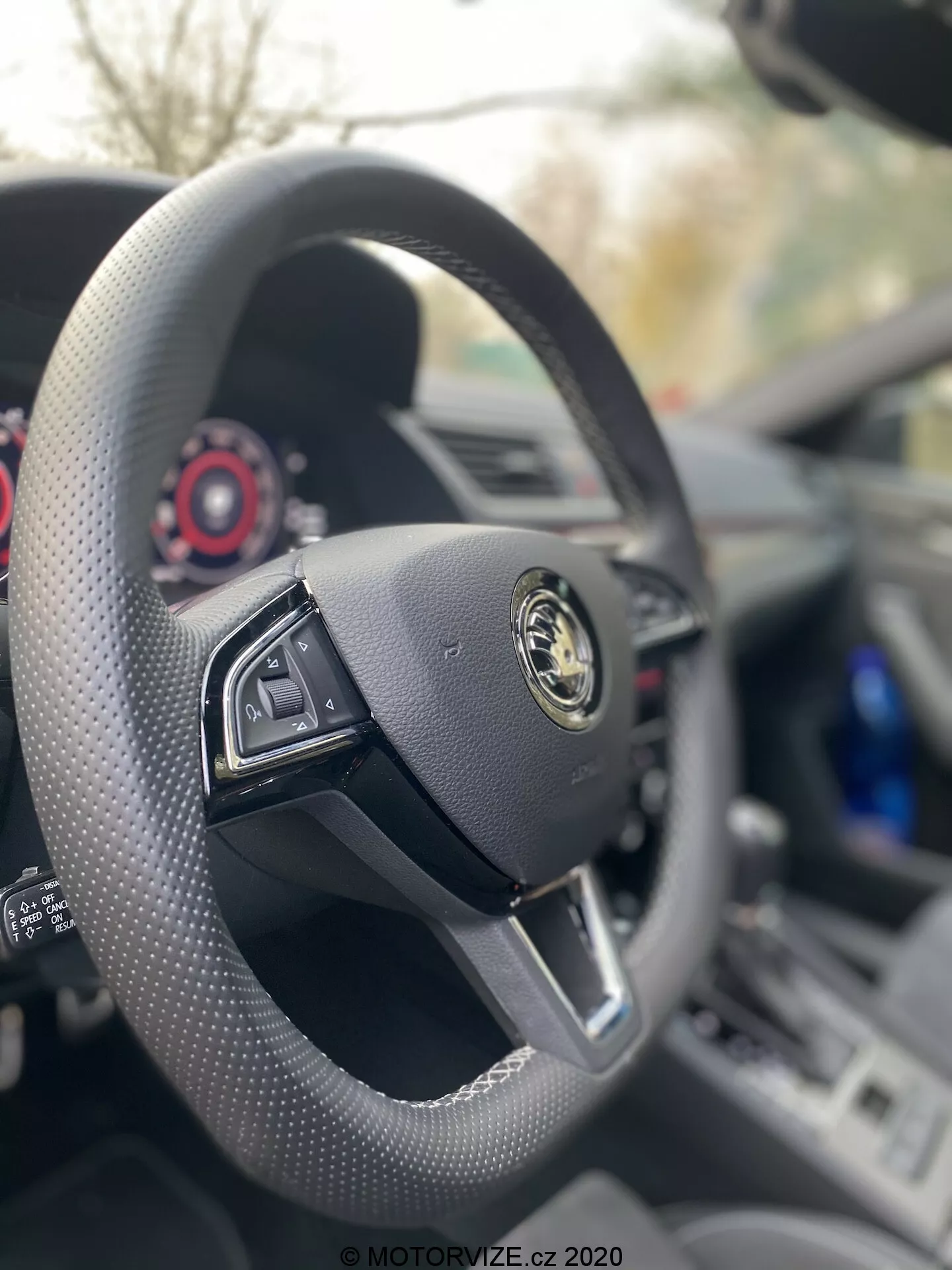 Pohľad na interiér Škody Superb Limousine 2019 z vodičovho sedadla, zameraný na čierny kožený volant s plochým spodkom a multifunkčnými ovládačmi. Logo výrobcu, so zlatým emblémom na čiernom pozadí, je výrazne viditeľné uprostred. Palubná doska a prístrojový panel sú viditeľné za volantom, zdôrazňujúc luxusný minimalistický dizajn vozidla s červenými detailmi na súčasných číselníkoch.