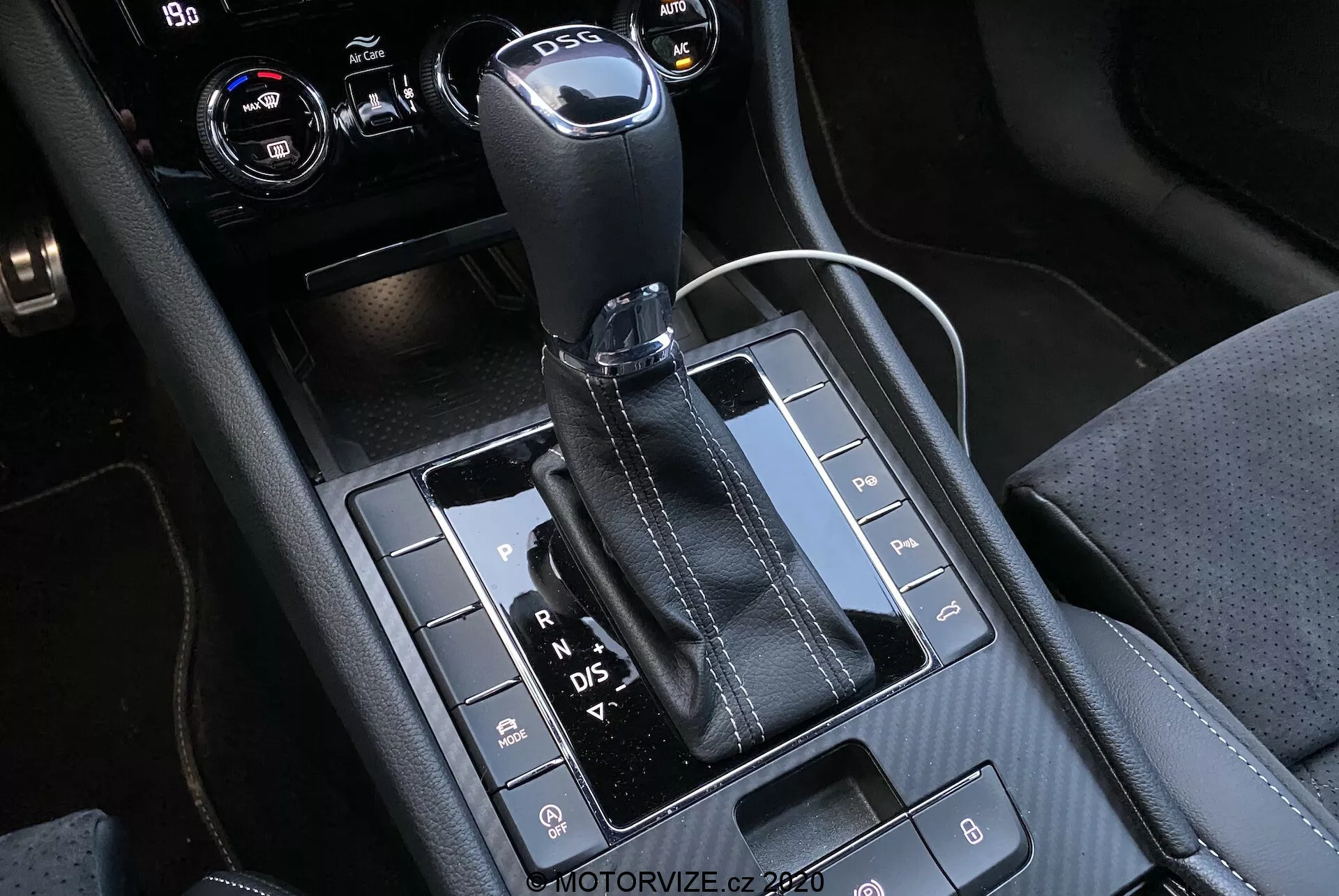 Pohľad na automatickú radiacu páku a okolitú stredovú konzolu automobilu Skoda Superb 2019, s koženou a kovovo zakončenou radiacou pákou s viditeľnými písmenami 'P, R, N, D/S', ktoré označujú rôzne stupne. Lesklý čierny panel obsahuje rôzne tlačidlá na funkcie ako parkovacie senzory, deaktiváciu systému Auto Start/Stop a kontrolu trakcie. Konzola obsahuje kombináciu materiálov s detailným šitím, vrátane 12-voltovej zásuvky a pravdepodobne ovládače klimatizácie alebo infotainmentu vo vyrezanom pravom hornom rohu.