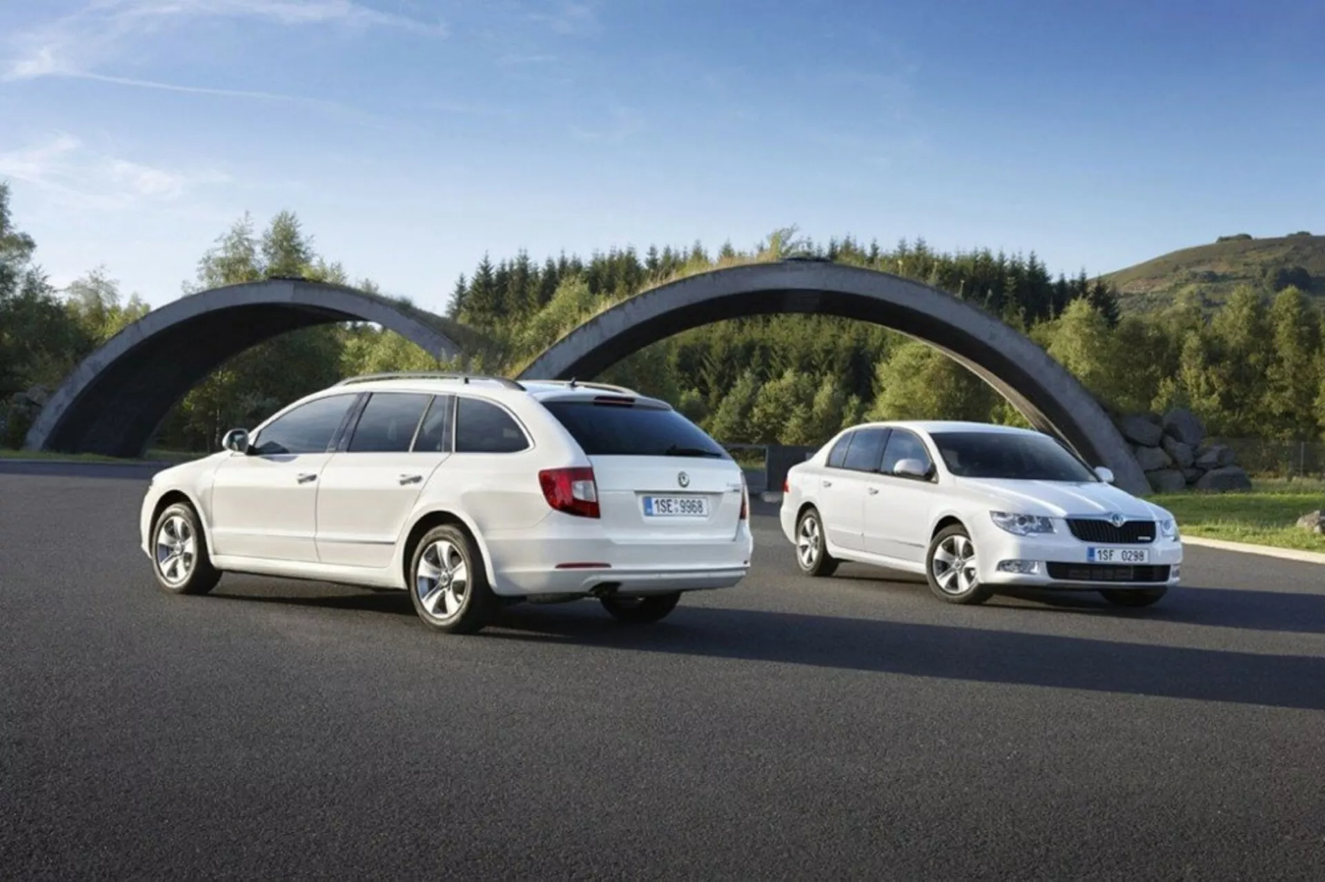 Škoda Superb kombi z roku 2008 vo dne, zobrazujúca zadné a predné tri štvrtiny uhlov na asfaltovej ceste proti zelene a zakrivenému mostu, zameriava sa na dizajnové prvky ako sú zadné svetlá, predná mriežka a silueta vozidla.