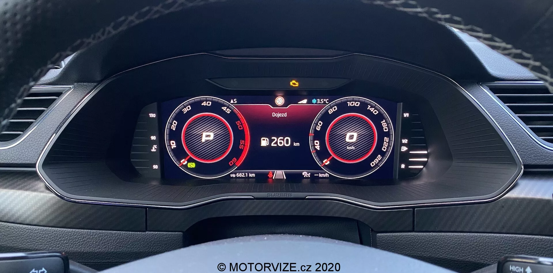 Pohľad do interiéru vozidla Škoda Superb Limousine 2019 so zameraním na plne digitálnu, vysokorozlišovaciu prístrojovú dosku z pohľadu vodiča. Displej zahŕňa dva veľké virtuálne merače rýchlomera a otáčkomera, osvetlené červenou a bielou farbou. Medzi meračmi sa zobrazujú ďalšie informácie, ako je ukazovateľ prevodového stupňa 'P', údaj o teplote a výstražný symbol. Ukazovateľ batérie naznačuje elektrické alebo hybridné schopnosti. Dizajn palubnej dosky zahŕňa vzduchové ventily a vrchnú časť tvarovaného volantu so šitými detailmi.