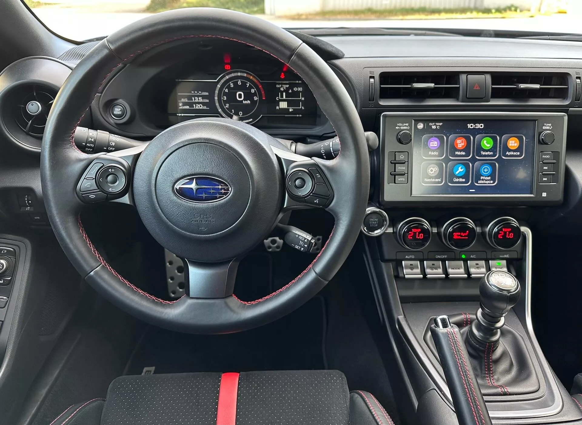Interiér Subaru BRZ 2023 z pohľadu vodiča zahŕňajúci športový kožený volant s multifunkčnými ovládacími prvkami a červeným stehom, prístrojový panel s analógovými ciferníkmi a digitálnym displejom, stredovú konzolu s veľkým dotykovým obrazovkou infotainment systému a dvojzónovú klimatizáciu, a manuálnu prevodovku s koženým čalúnením a červenými akcentmi, na pozadí čierneho interiéru so športovými červenými akcentmi, zdôrazňujúci pokročilé funkcie vozidla a technológiu.