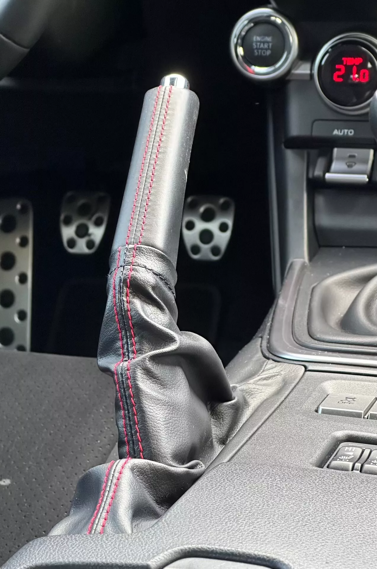 Detailný pohľad na stredovú konzolu Subaru BRZ 2023 s dôrazom na kovovú radicu páku s červeným kontrastným šitím na koženom čermeďu, digitálne zobrazenie '21', športové hliníkové pedále s vrtanými otvormi a technologické funkcie ako tlačidlá pre elektronickú stabilitu.