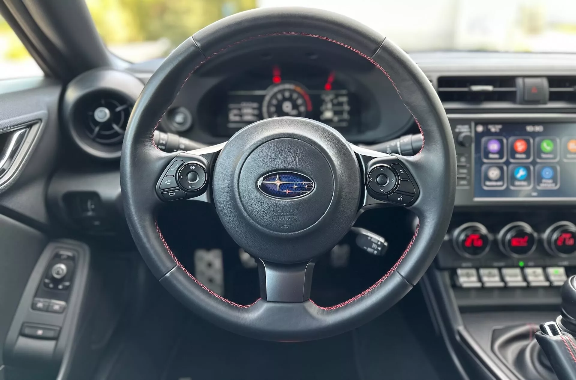 Interiér Subaru BRZ 2023 z pohľadu vodiča zameraný na trojramenný volant s červeným šitím, logo Subaru a palubnú dosku s prístrojovým panelom a stredovou konzolou