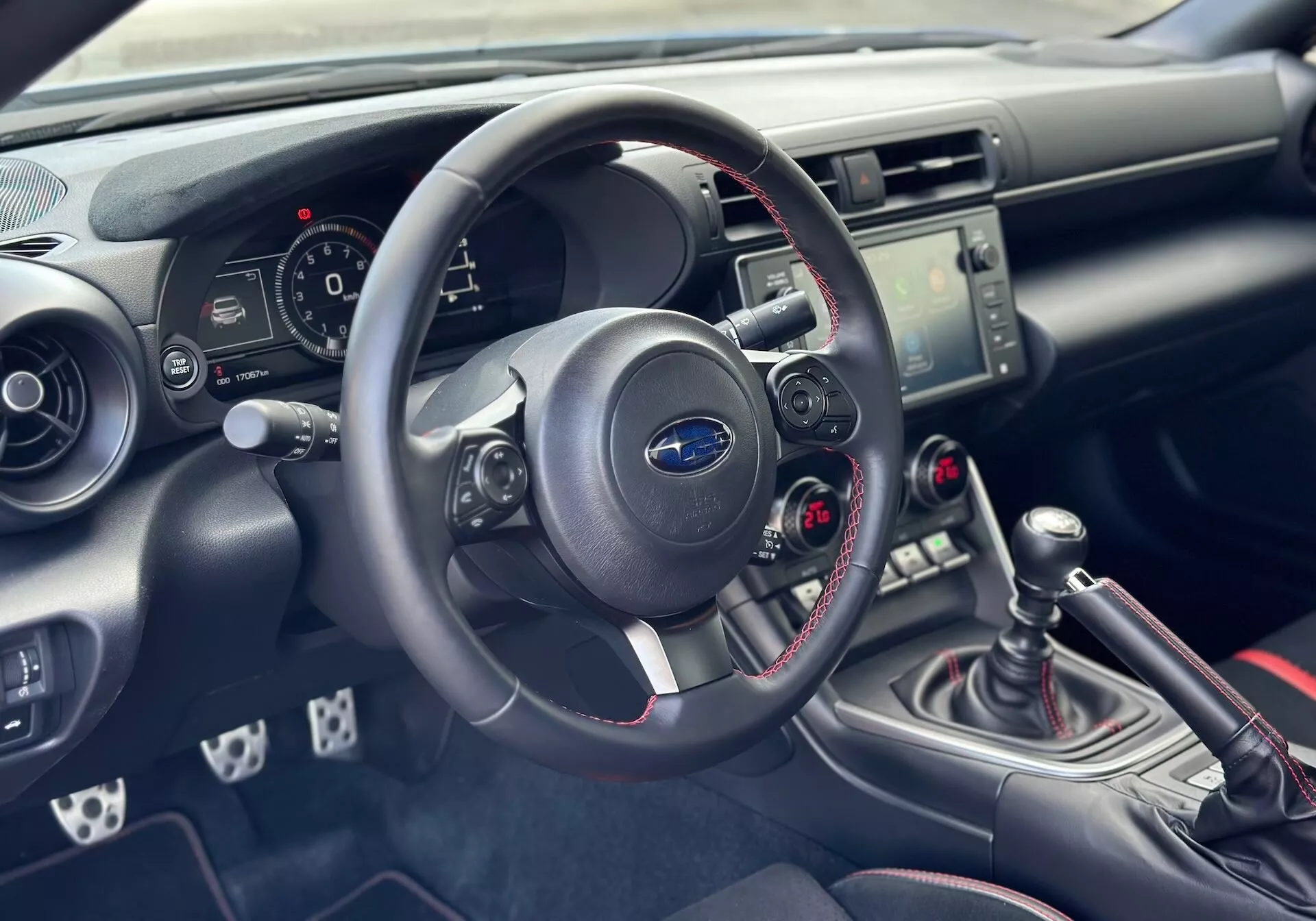 Interiér Subaru BRZ 2023 so zameraním na palubnú dosku a kokpit na strane vodiča s detailným pohľadom na volant, prístrojový panel, stredovú konzolu s ovládaním audia a klímy, a manuálnu radiacu páku so športovým červeným prešívaním
