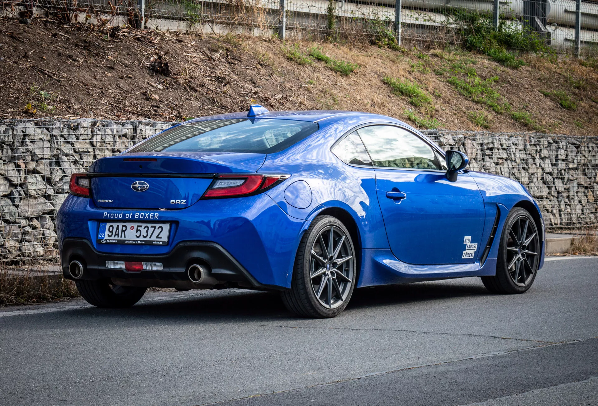 Športové kupé Subaru BRZ 2023 v metalickej modrej farbe zaparkované na svahu, ktoré zdôrazňuje zadný trojštvrtečný pohľad vozidla, bočný profil, zakrivenie zadnej časti, športové zliatinové kolesá, dvojité výfukové koncovky, malý spoiler na veku batožinového priestoru a odznak motora 'Proud of Boxer'.
