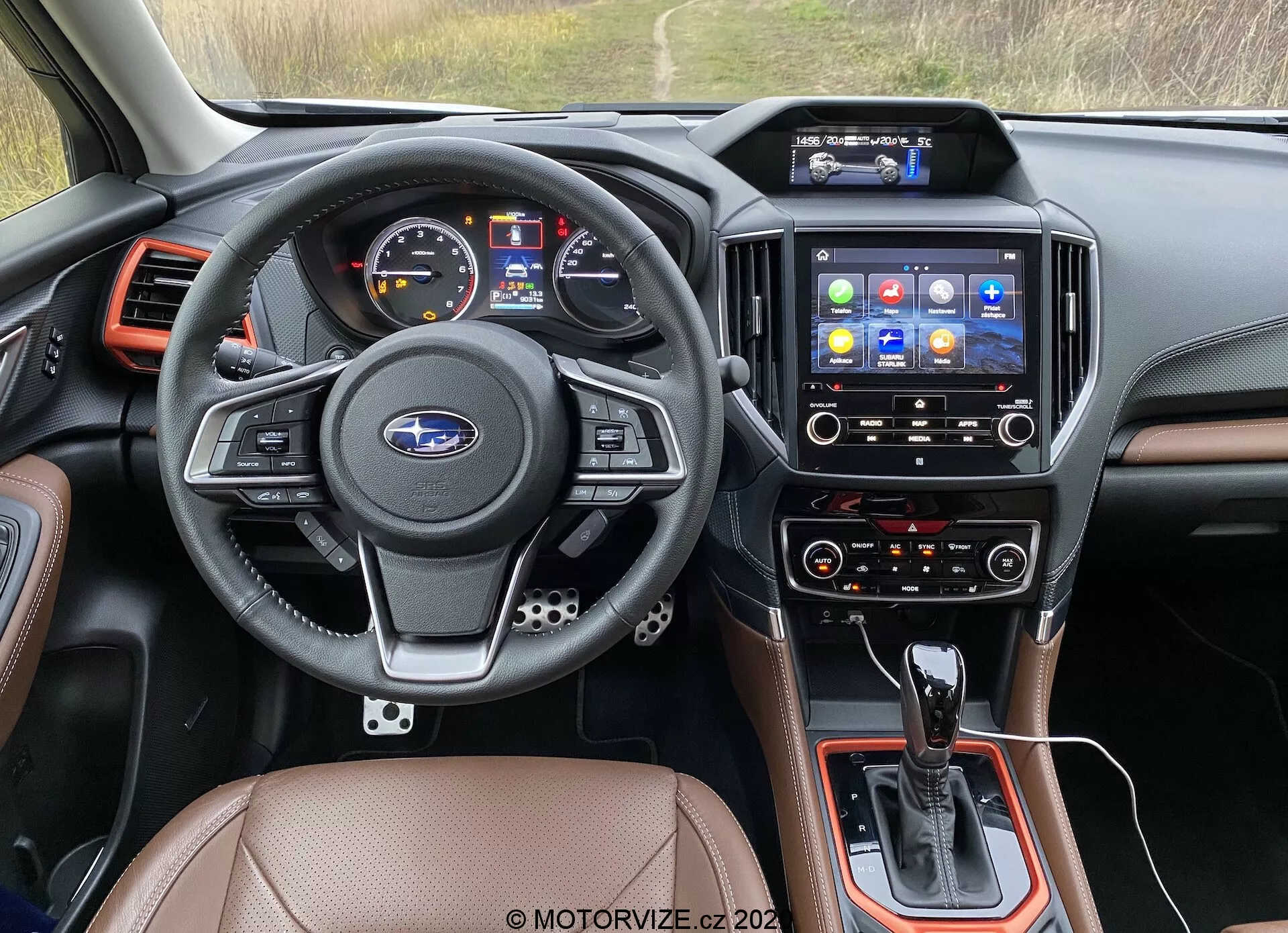 Záber interiéru z pohľadu vodiča modelu Subaru Forester 2020, so zameraním na palubnú dosku, volant, infotainment systém a stredovú konzolu. Trojramenný kožený volant ponúka multifunkčné ovládanie audia, telefonovania, hlasových príkazov a tempomatu, s emblémom Subaru uprostred. Prístrojový štít zahŕňa digitálny displej a analógové ukazovatele rýchlosti a otáčok. Stredová konzola obsahuje radiacu páku automatickej prevodovky, ďalšie ovládacie prvky a možný volič jazdných režimov. Infotainment dotyková obrazovka s rôznymi ikonami aplikácií ponúka navigáciu, médiá, telefónnu konektivitu a nastavenia vozidla. Nižšie sú ovládacie prvky klimatizácie, ktoré naznačujú dvojzónové nastavenie pre vodiča a predného spolujazdca. Interiér je upravený koženými alebo koženkovými sedadlami, čiernymi plastovými komponentmi a kovovými akcentmi. Vzduchové výduchy sú umiestnené okolo infotainment systému a na okrajoch palubnej dosky. Tento obrázok patrí do kategórií interiéry áut, automobilová technológia, palubné dosky, infotainment systémy a ovládanie klimatizácie.
