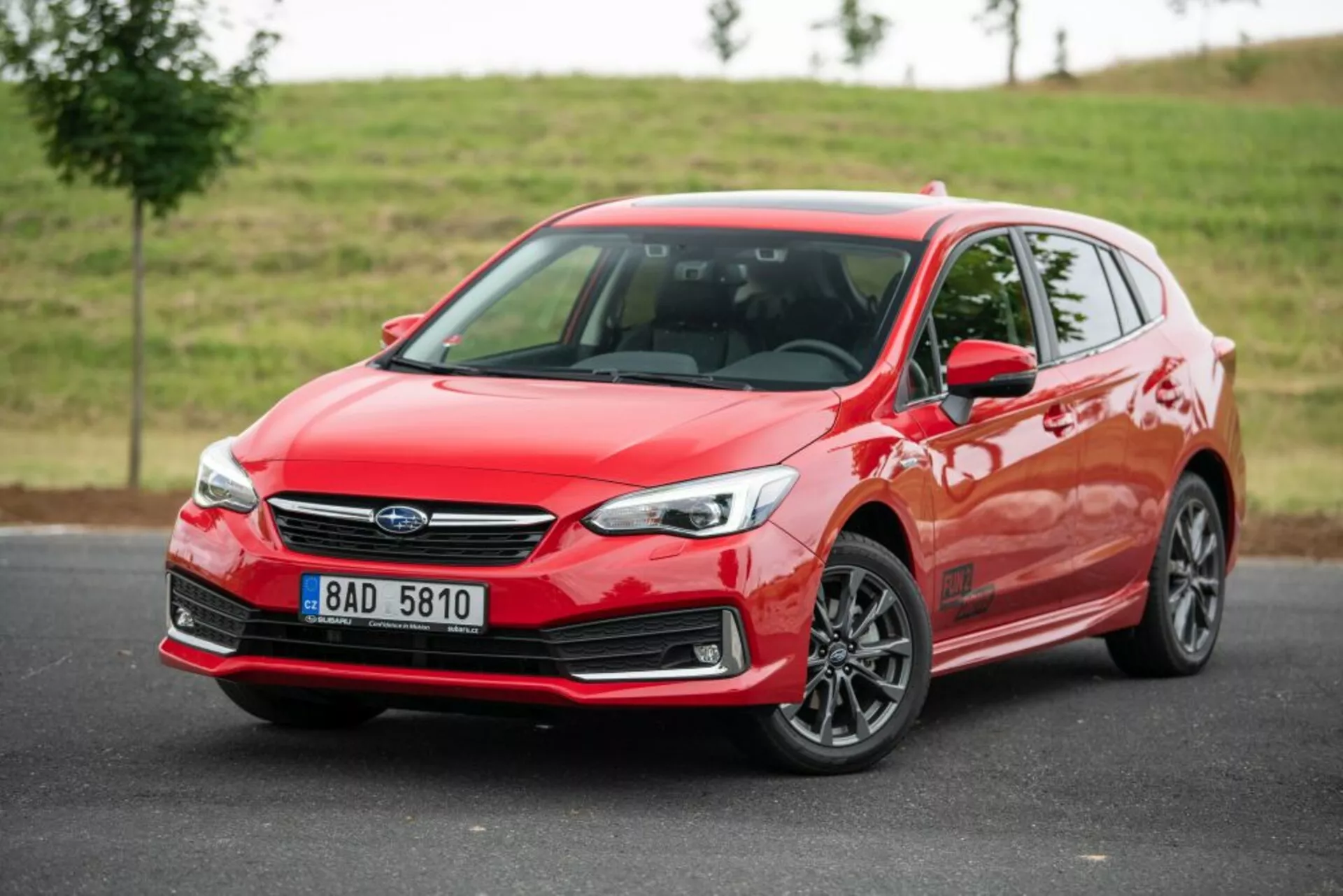 Svetlo červený hatchback Subaru Impreza 2018 s trojštvrtečným predným pohľadom, zobrazujúci jeho štíhle LED svetlomety, charakteristickú mriežku s centrálnym emblémom a štýlové viacprúdové zliatinové kolesá proti zamračenému, prírodnému pozadiu