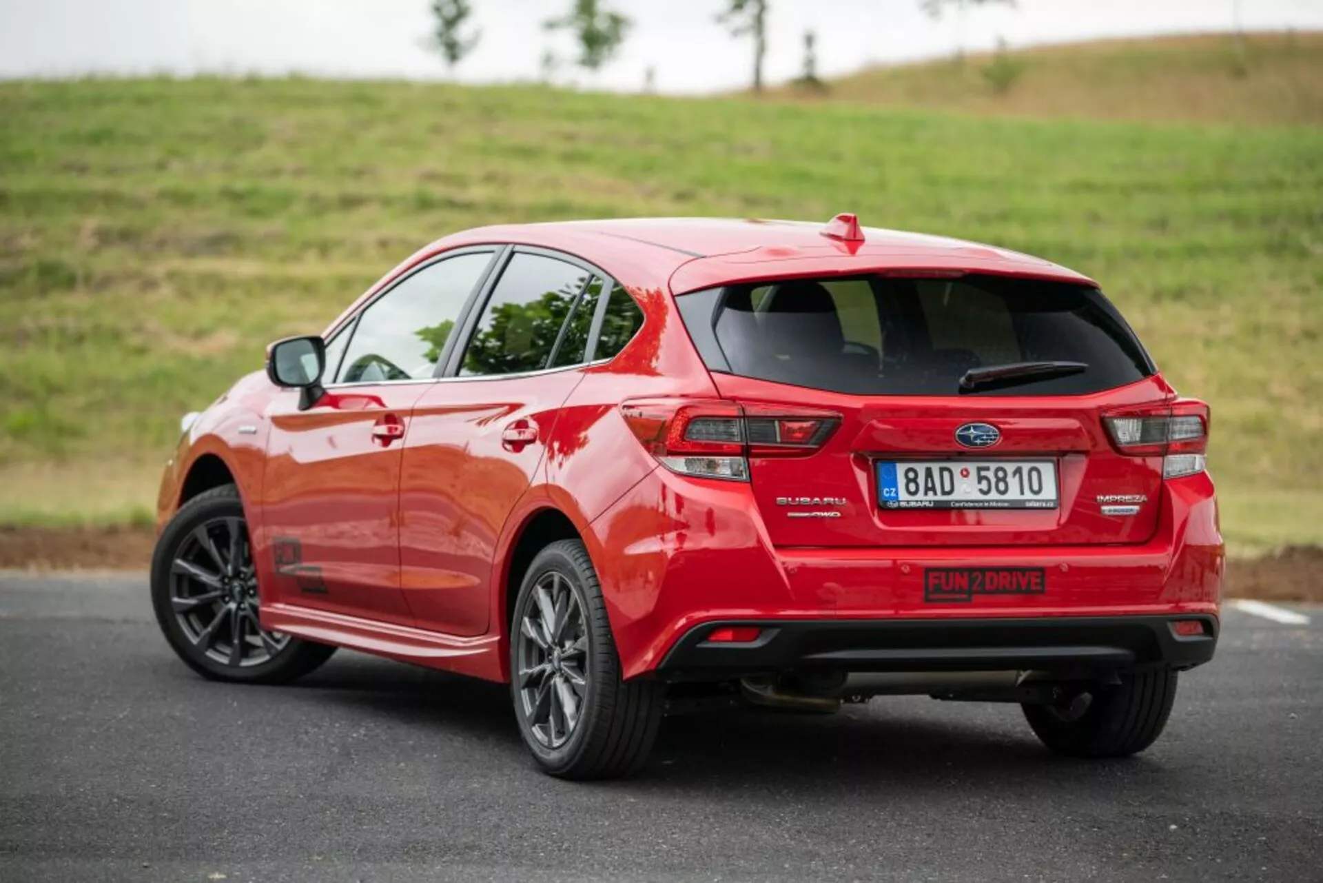 Červený hatchback Subaru Impreza z roku 2018 zobrazený z zadného trojštvrtečného uhla, ktorý zdôrazňuje jeho bočný profil, charakteristický dizajn zadných svetiel, výfukový systém, spoiler a zliatinové kolesá na asfaltovej ceste s trávnatým pozadím pri mäkkom zamračenom osvetlení