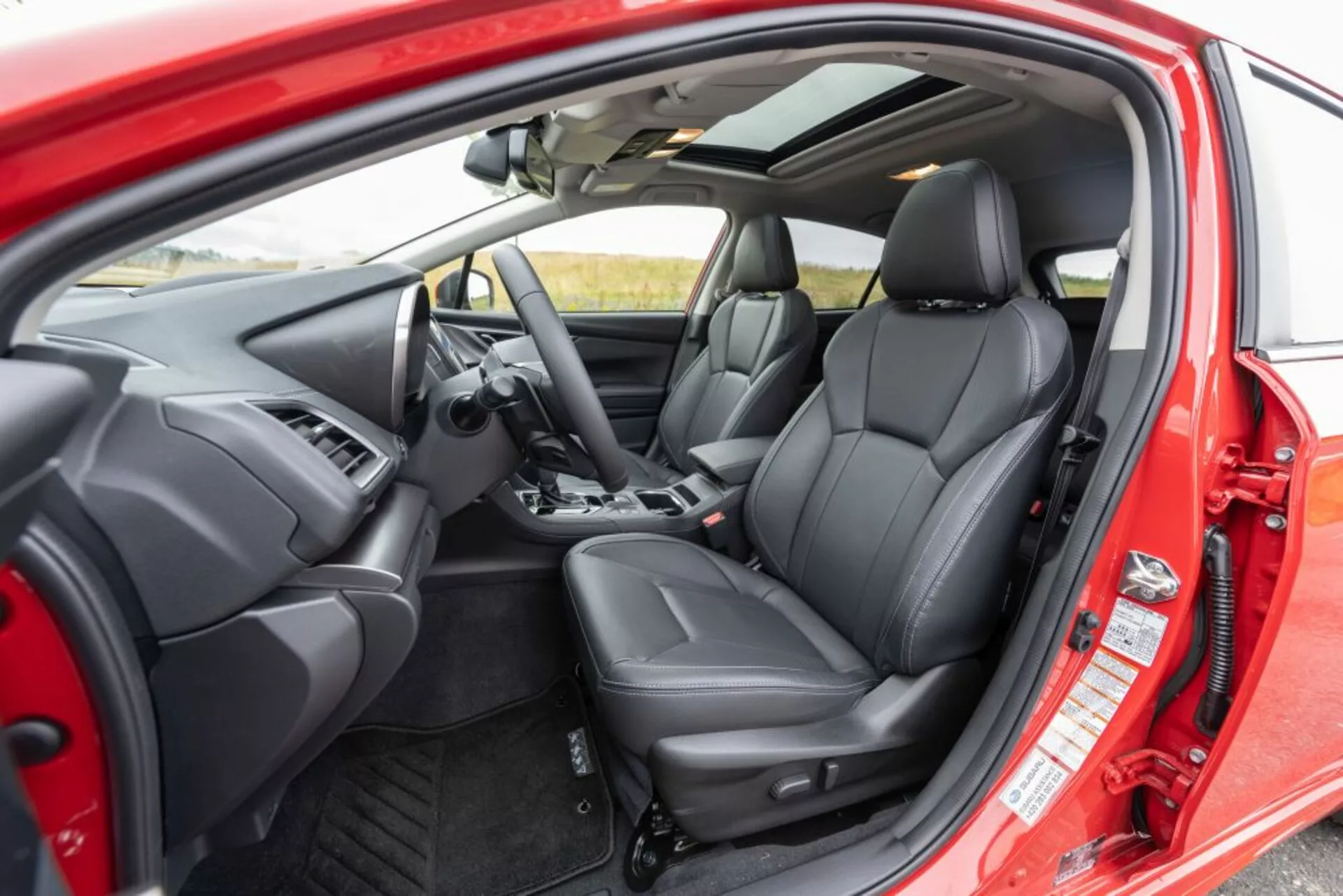 Interiér Subaru Impreza 2018 z vonkajšej perspektívy ukazuje sedadlo vodiča, prístrojovú dosku, volant s ovládacími tlačidlami, čalúnené predné sedadlá v čiernej koži s ergonomickým dizajnom a ovládanie na vnútornej strane dverí cez otvorené dvere vodiča