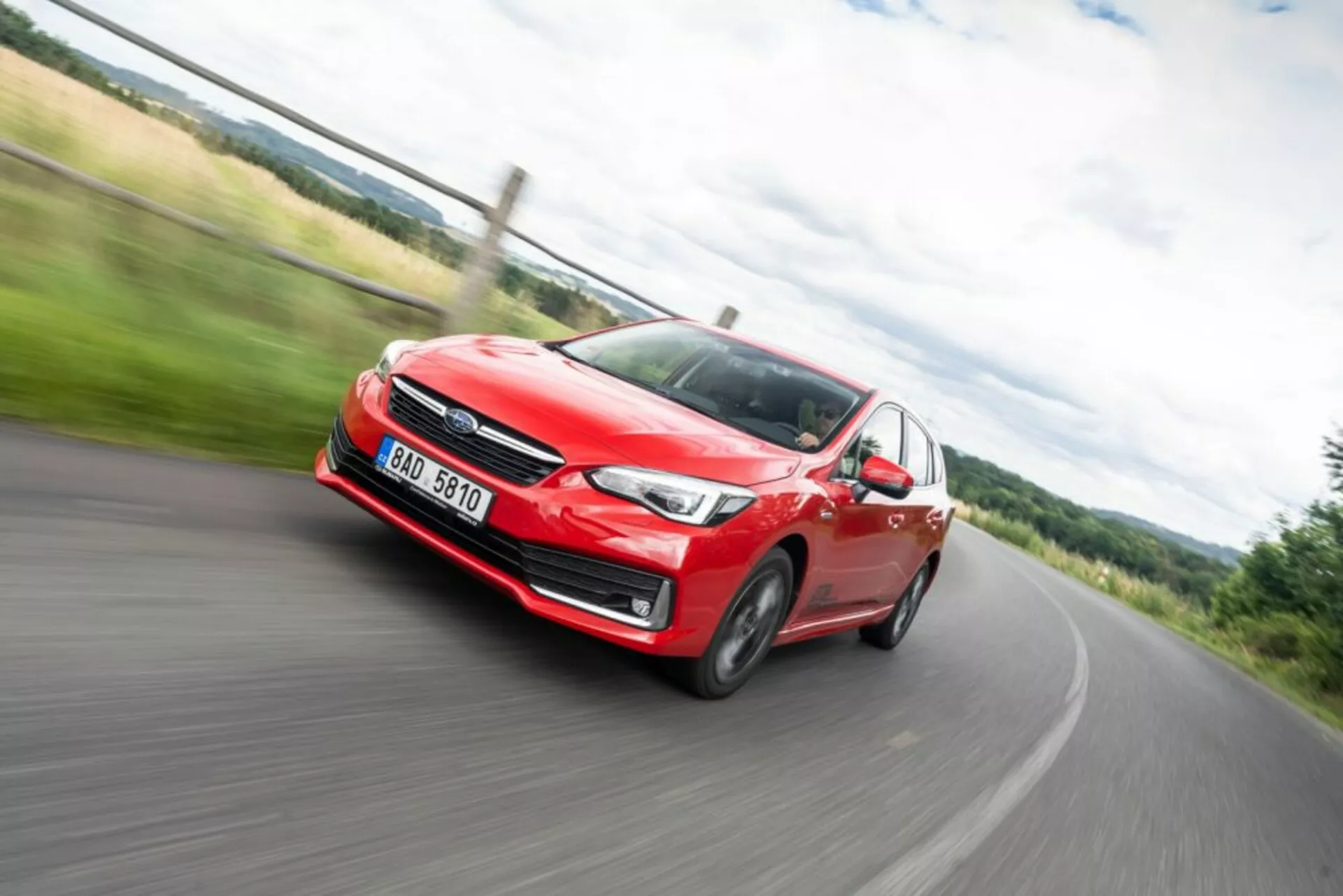 Dynamický obrázok s rozmazaním pohybu červeného Subaru Impreza z roku 2018 v akcii, pohľad spredu ukazujúci svetlomet, mriežku a zliatinové kolesá počas zatáčania vľavo na vidieckej ceste