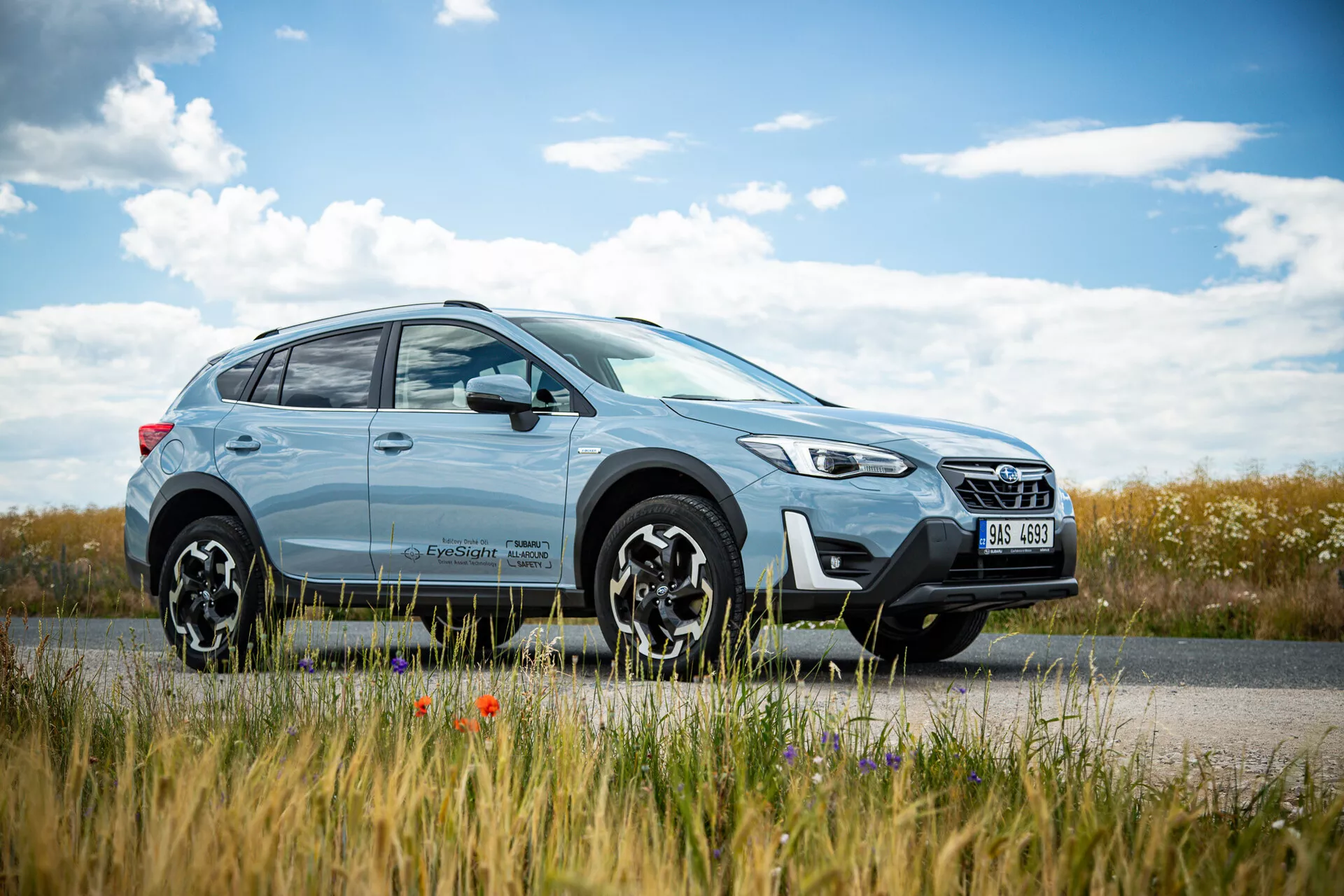 Svetlomodrý crossover SUV Subaru XV 2021 zaparkovaný pri ceste so scenériou vidieka na pozadí, predstavujúci moderný dizajn s LED svetlometmi a zliatinovými kolesami s viacerými lúčmi, zdôrazňujúci schopnosti jazdy v meste aj v teréne.