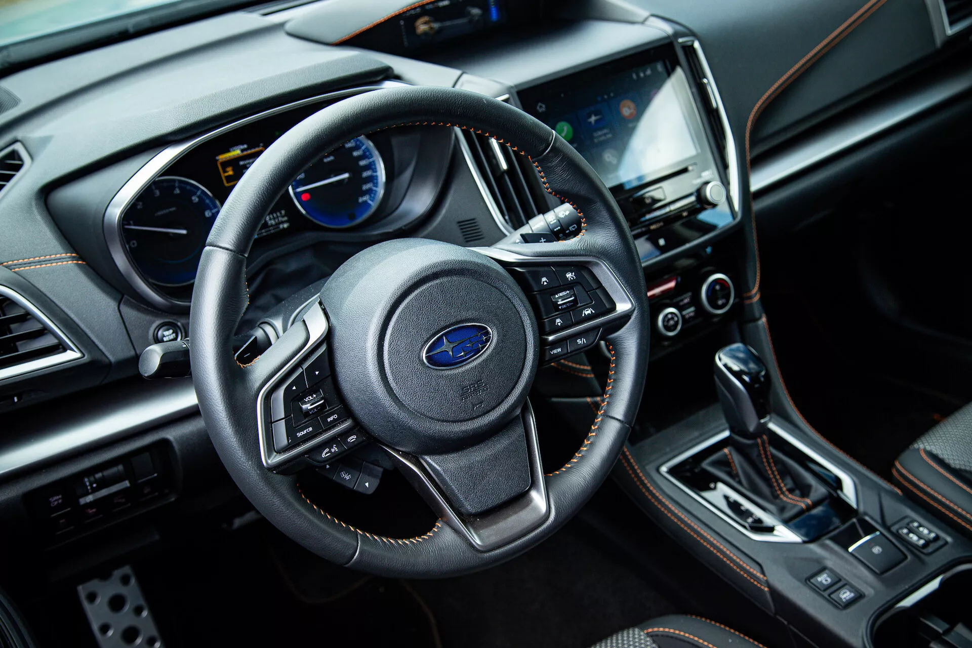 Interiér Subaru XV 2021 z pohľadu predného spolujazdca ukazujúci trojramenný volant s multifunkčnými ovládačmi, prístrojový panel s analógovými a digitálnymi displejmi na palubnej doske, centrálny dotykový infotainment systém, tlačidlá klimatizácie, páku automatickej prevodovky a luxusné materiály, vrátane kože a chrómových akcentov.