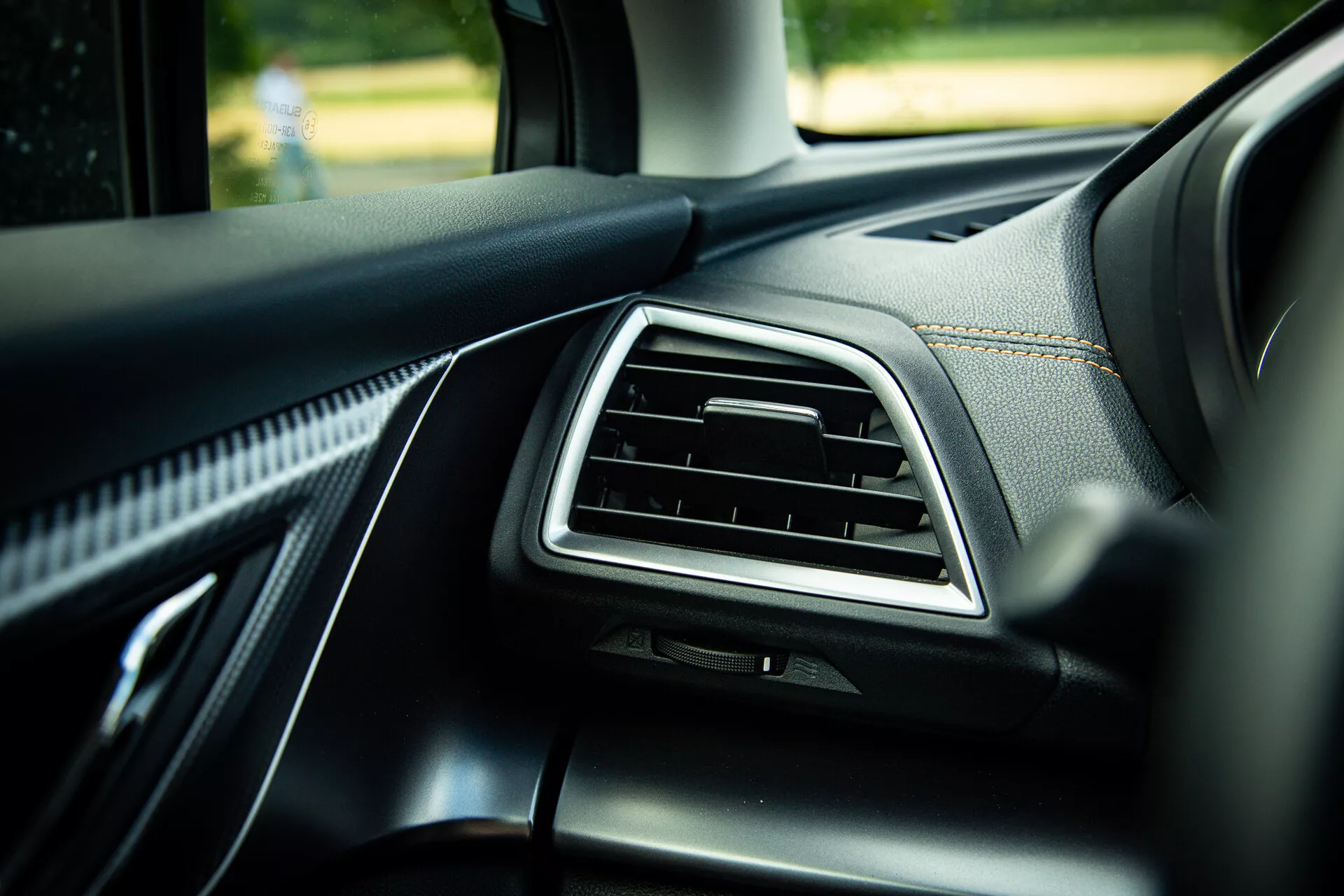 Detail interiéru Subaru XV 2021 zobrazujúci palubnú dosku na strane spolujazdca s vetracím otvorom zdôrazneným chrómom, kvalitným čiernym koženým čalúnením so zreteľným šitím a materiálmi príjemnými na dotyk, fotografovaný pod uhľom 45 stupňov, ktorý zvýrazňuje moderné dizajnové prvky pri prirodzenom dennom svetle