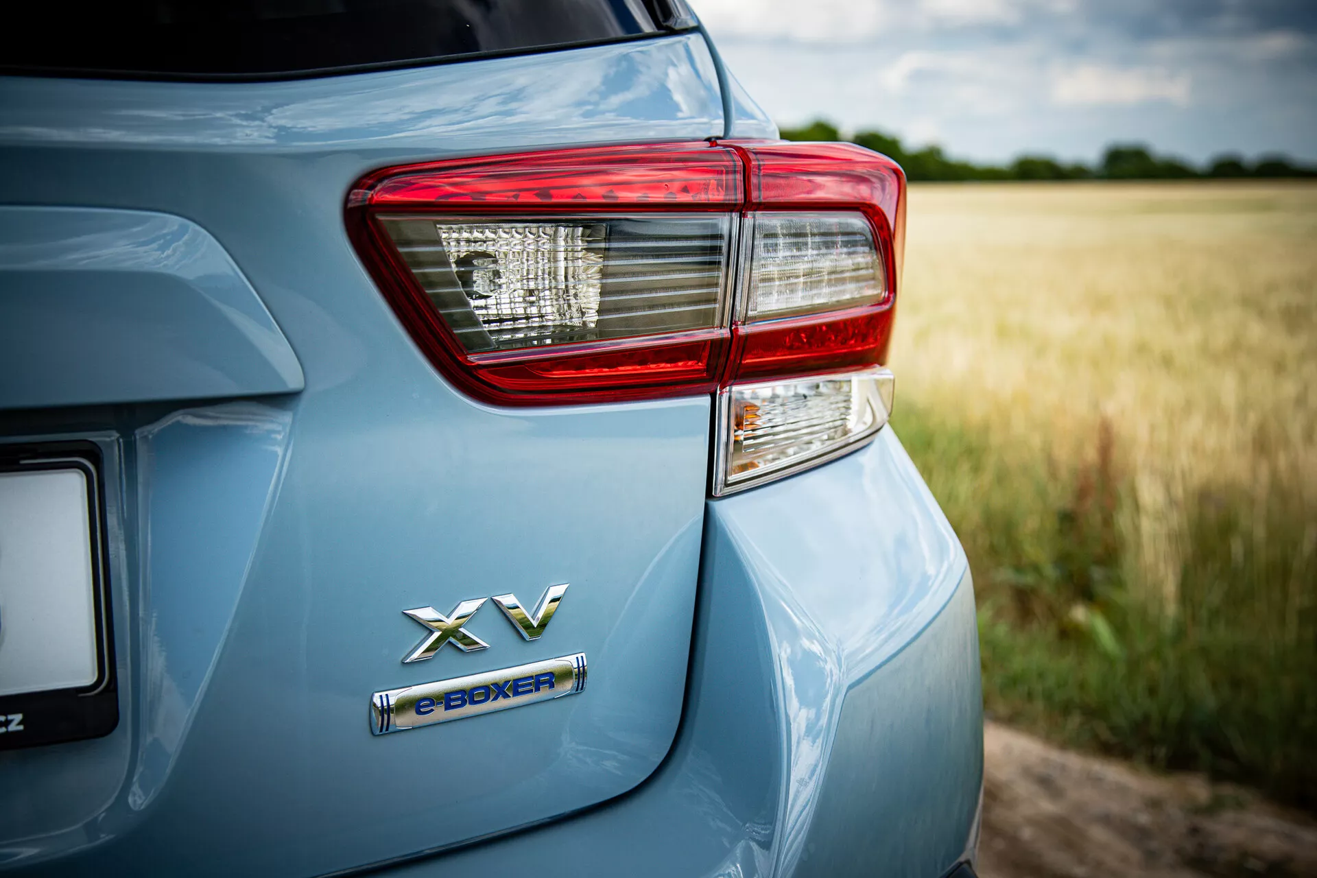 Detailný pohľad na zadnú ľavú stranu Subaru XV 2021 s emblémom hybridného pohonu e-BOXER a podrobne spracovaným svetlom zadného svetla na metalickom svetlomodrom laku v vidieckom prostredí