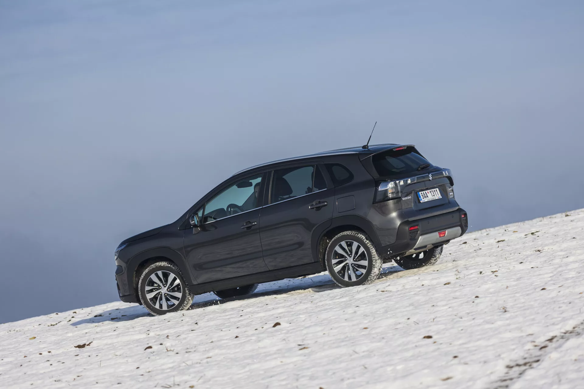 SUV Suzuki S-Cross 2022 v tmavom odtieni umiestnené na zasneženom svahu zachytené z šikmého zadného uhla zvýrazňujúce jeho bočný a zadný profil, veľké zliatinové kolesá, tónované okná s odmrazovačom, LED zadné svetlá, strešný spoiler a potenciálne off-road schopnosti pod podmienkami jasného neba.