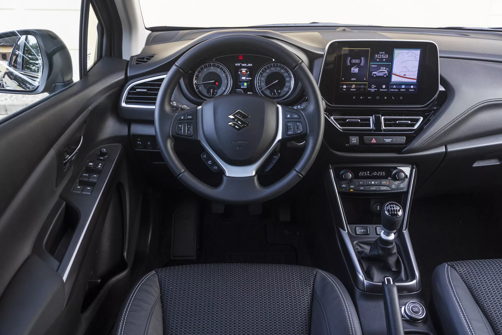 Interiér prednej kabíny Suzuki S-Cross 2022 zobrazujúci prístrojovú dosku, volant s ovládacími prvkami, digitálno-analógový kombinovaný prístrojový panel, dotykový infotainment systém, ovládače klímy, koženú páku radenia, volič jazdného režimu, elektronickú parkovaciu brzdu a dverový panel s tlačidlami ovládania
