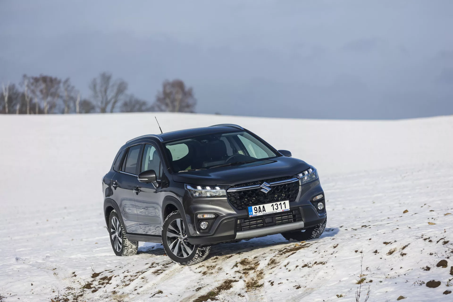 Tmavé kompaktné SUV Suzuki S-Cross ročník 2022 zaparkované v zasneženej krajine, ukazujúce pohľad zpredu a z boku pod uhlom 45 stupňov, s elegantnou mriežkou s kovovým povrchom, predĺženými LED svetlometmi, zliatinovými kolesami s viacerými lúčmi a lešteným chrómovým olemovaním okien naznačujúcim luxusnú výbavu a potenciálne schopnosť terénnej jazdy