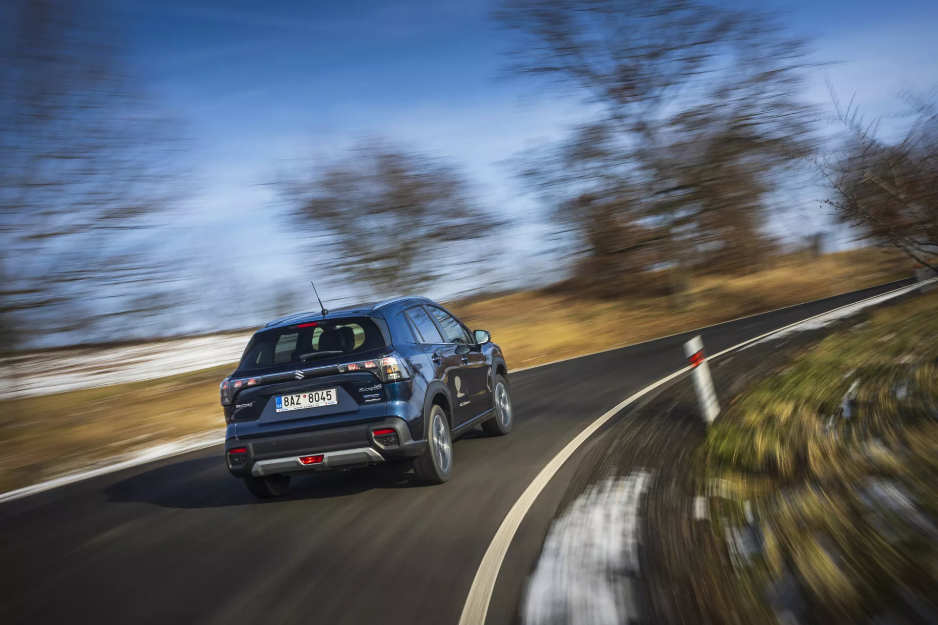 Moderné SUV Suzuki S-Cross 2022 v pohybe, ukazuje profil z pohľadu zadnej trojštvrtečnej časti s rotujúcimi kolesami na jasný deň