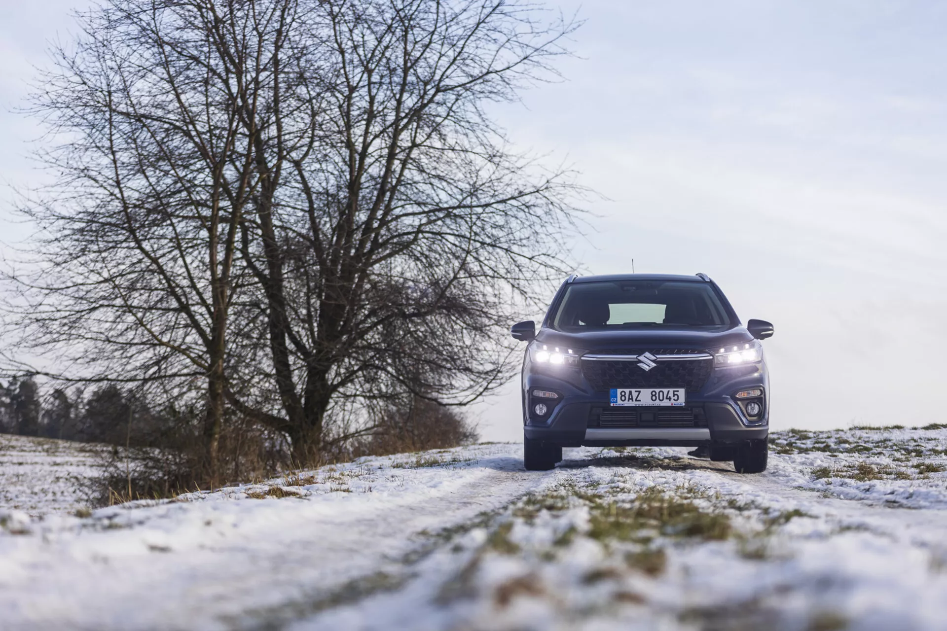 Predný pohľad na Suzuki S-Cross 2022 na zasneženej vidieckej ceste, predvádzajúci LED svetlomety, moderný dizajn mriežky, zliatinové kolesá a elegantnú karosériu SUV na pozadí zimy so stromami bez listov a zamračnou oblohou.