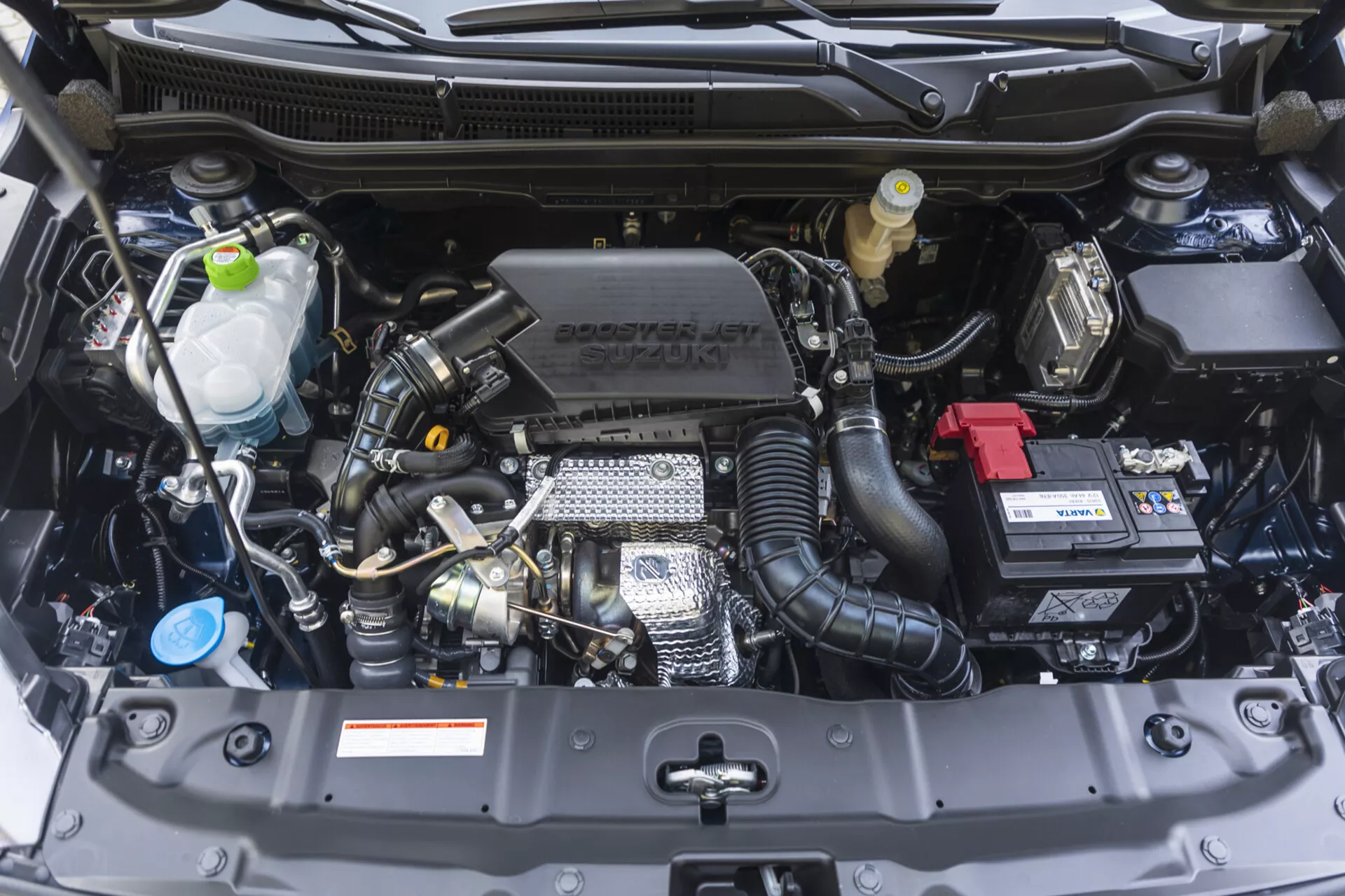 Pohľad zpredu na motorový priestor Suzuki S-Cross 2022 s viditeľnými komponentmi ako sú blok motora, hlava valca, sací potrubí a technologickými prvkami vrátane turbodúchadla a variabilného časovania ventilov, spolu so značením Suzuki na kryte motora, v bezchybnom stave.