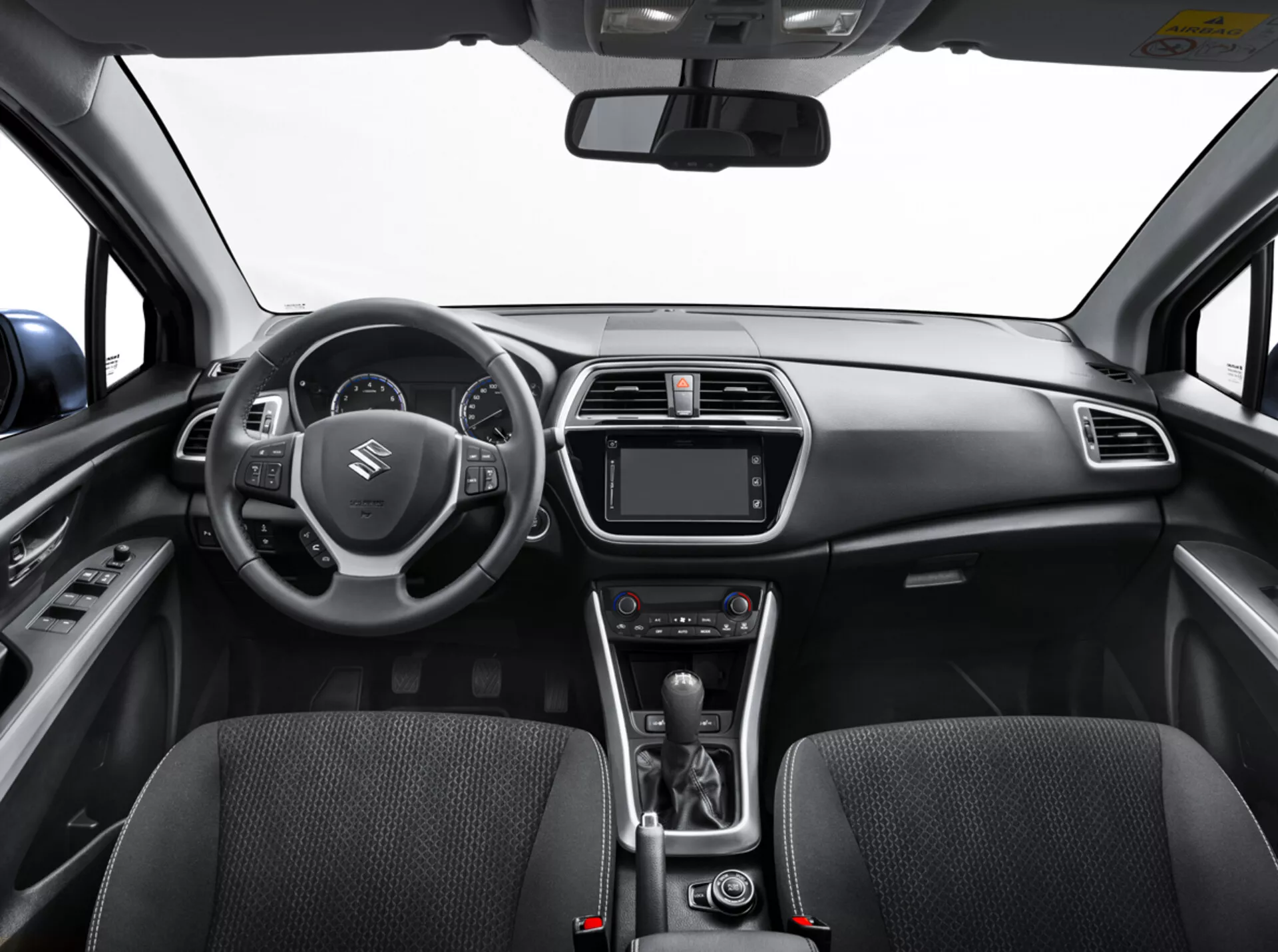 Interiér Suzuki S-Cross 2022 z pohľadu zadného sedadla ukazujúci palubnú dosku, volant s ovládacími prvkami, digitálny prístrojový panel, systém infotainmentu, ovládanie klímy, páku automatickej prevodovky a detailne spracované predné sedadlá s textúrou látky