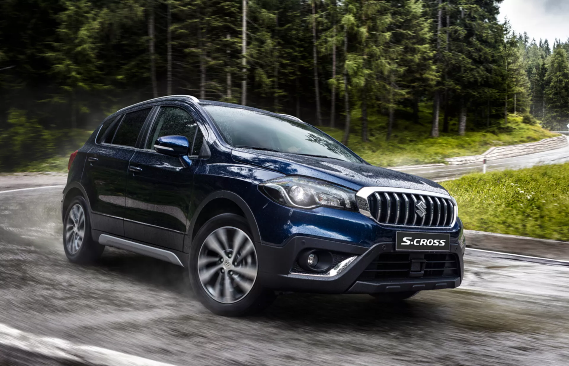 Suzuki S-Cross 2022 zachytený z predného trojštvrtečného uhla, zvýrazňujúci jeho mriežku s chromovanými akcentami, moderné LED alebo HID svetlomety, zliatinové kolesá s viacermi špicami a robustný crossoverový tvar tela na mokrej ceste a v zeleni, naznačujúci pokročilú technológiu a schopnosť jazdy za akéhokoľvek počasia.