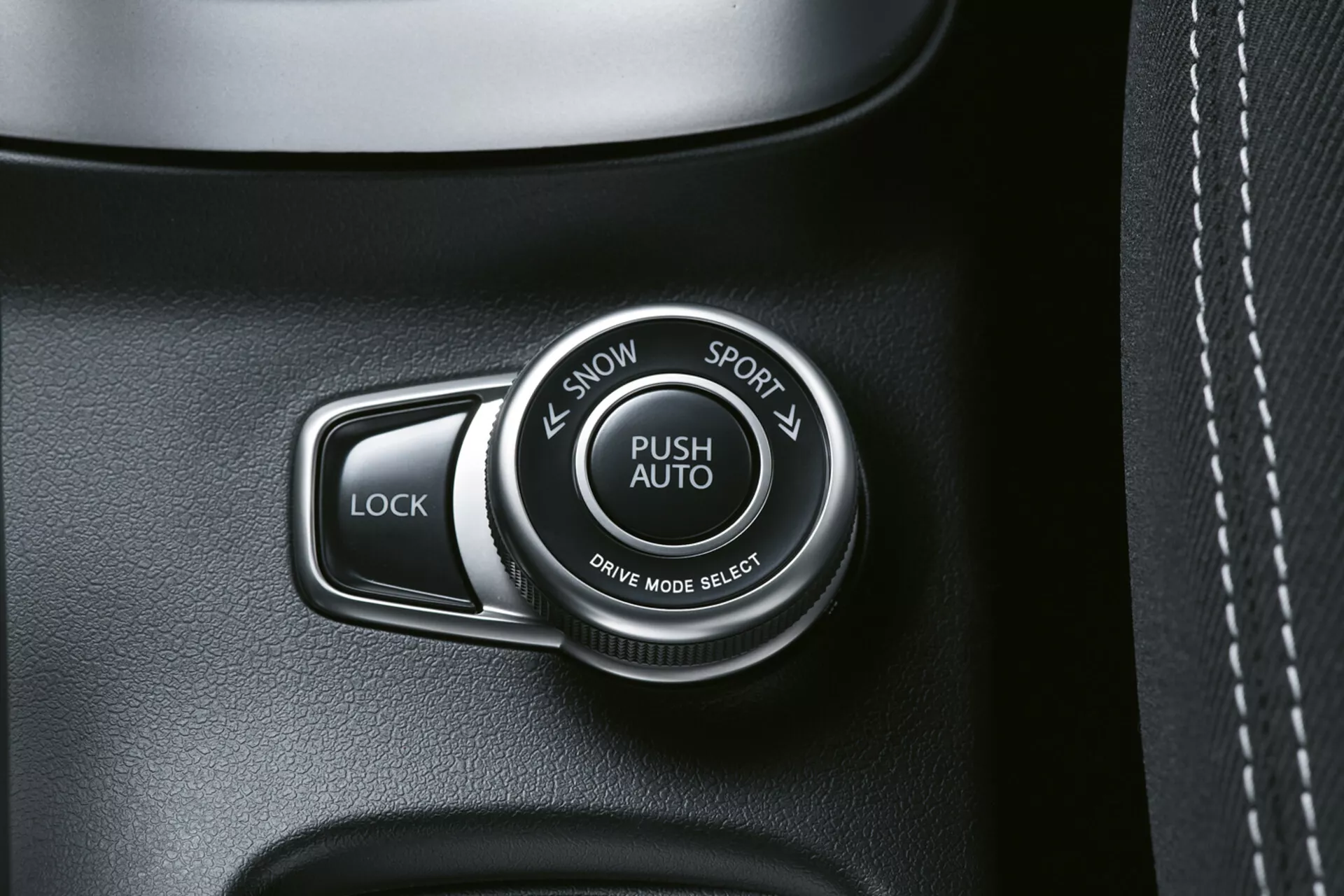 Detailný pohľad na interiér Suzuki S-Cross 2022 ukazujúci prepínač režimu jazdy označený 'PUSH AUTO' s možnosťami 'SNOW' a 'SPORT', vedľa tlačidla 'LOCK', na kvalitnom paneli tmavej farby, možno s koženou textúrou, s viditeľným šitím.