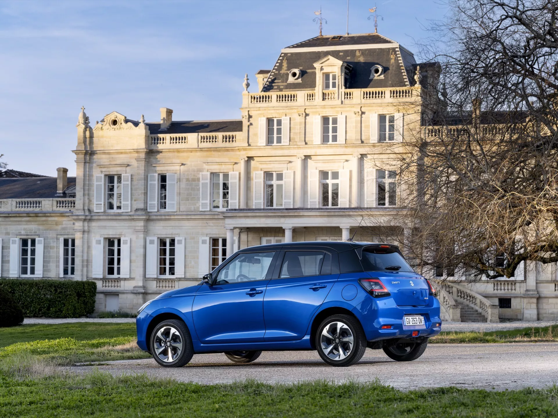 Suzuki Swift 7. generácia z roku 2024 v modrej farbe, parkovaný na štrkovej príjazdovej ceste pred budovou s klasickou architektúrou, ktorá zvýrazňuje dizajn vozidla z uhla tri štvrtiny zozadu s LED zadnými svetlami, zliatinovými kolesami a možnými modernými bezpečnostnými prvkami.