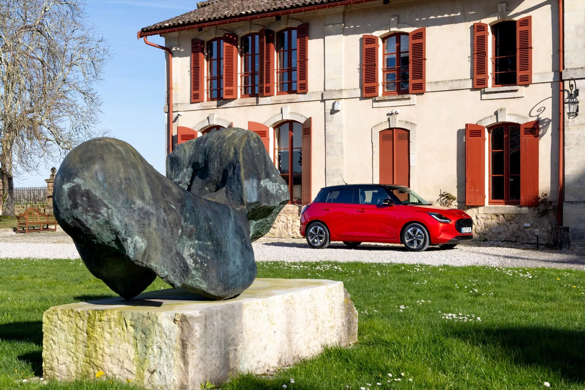 Suzuki Swift 7. generácie 2024 v žiarivo červenej farbe, zaparkované vonku, predvádzajúce jeho športový dizajn hatchbacku, zliatinové kolesá a tónované okná, s nápadným bočným pohľadom a perspektívou zadného štvrťroka, nastavené proti klasickému budovu a abstraktnú kameňovú sochu