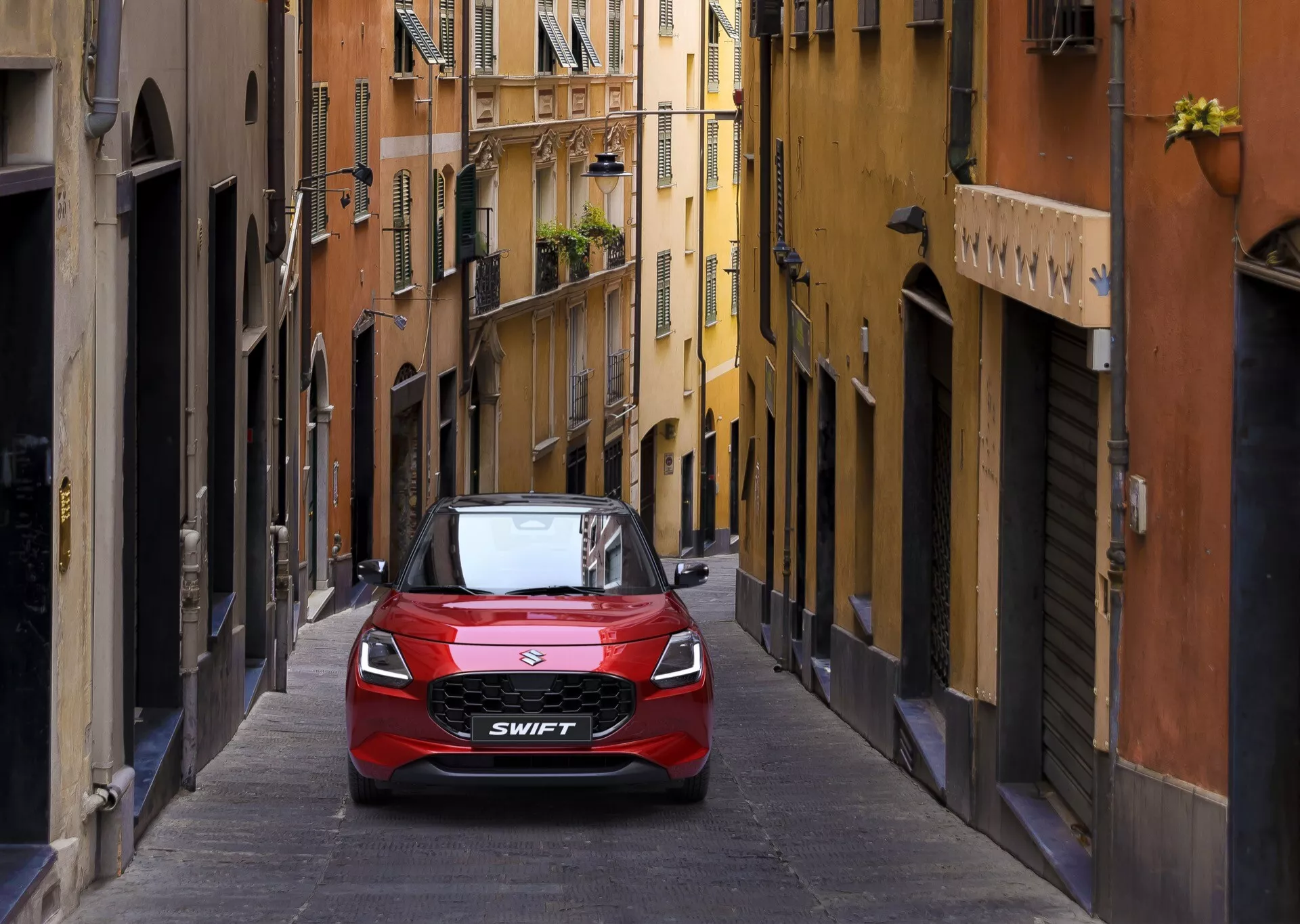 Červený Suzuki Swift 7. generácie z roku 2024 predstavený na malebnej európskej uličke, zdôrazňujúci jeho kompaktný dizajn a modernú prednú mriežku so značkovým logom, naznačujúci mestskú praktickosť a súčasný štýl.