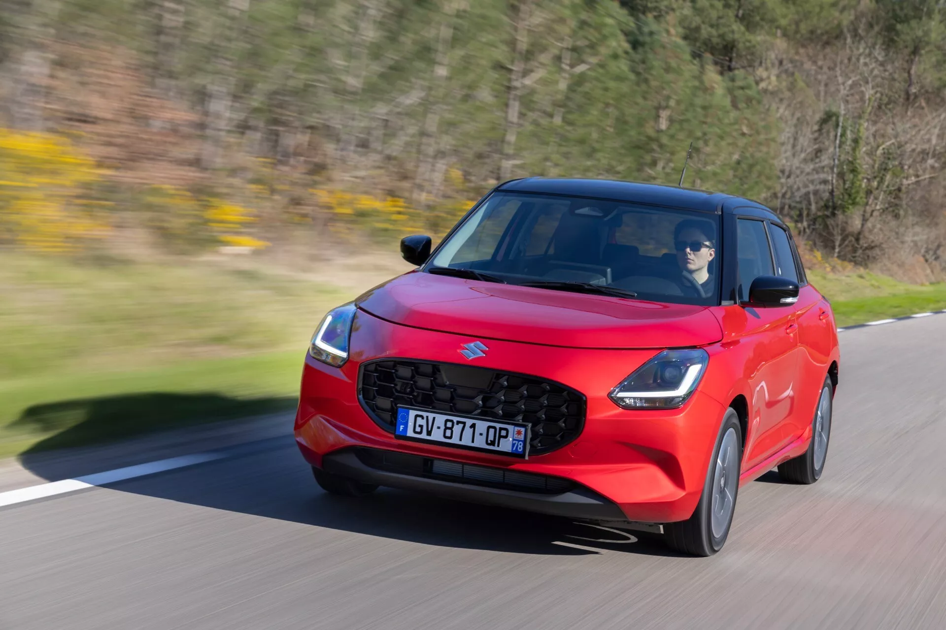 Suzuki Swift 2024 7. generácie v pohybe, červený hatchback s ostrým aerodynamickým dizajnom, LED svetlometmi a dynamickým stylingom proti rozmazanej zeleni