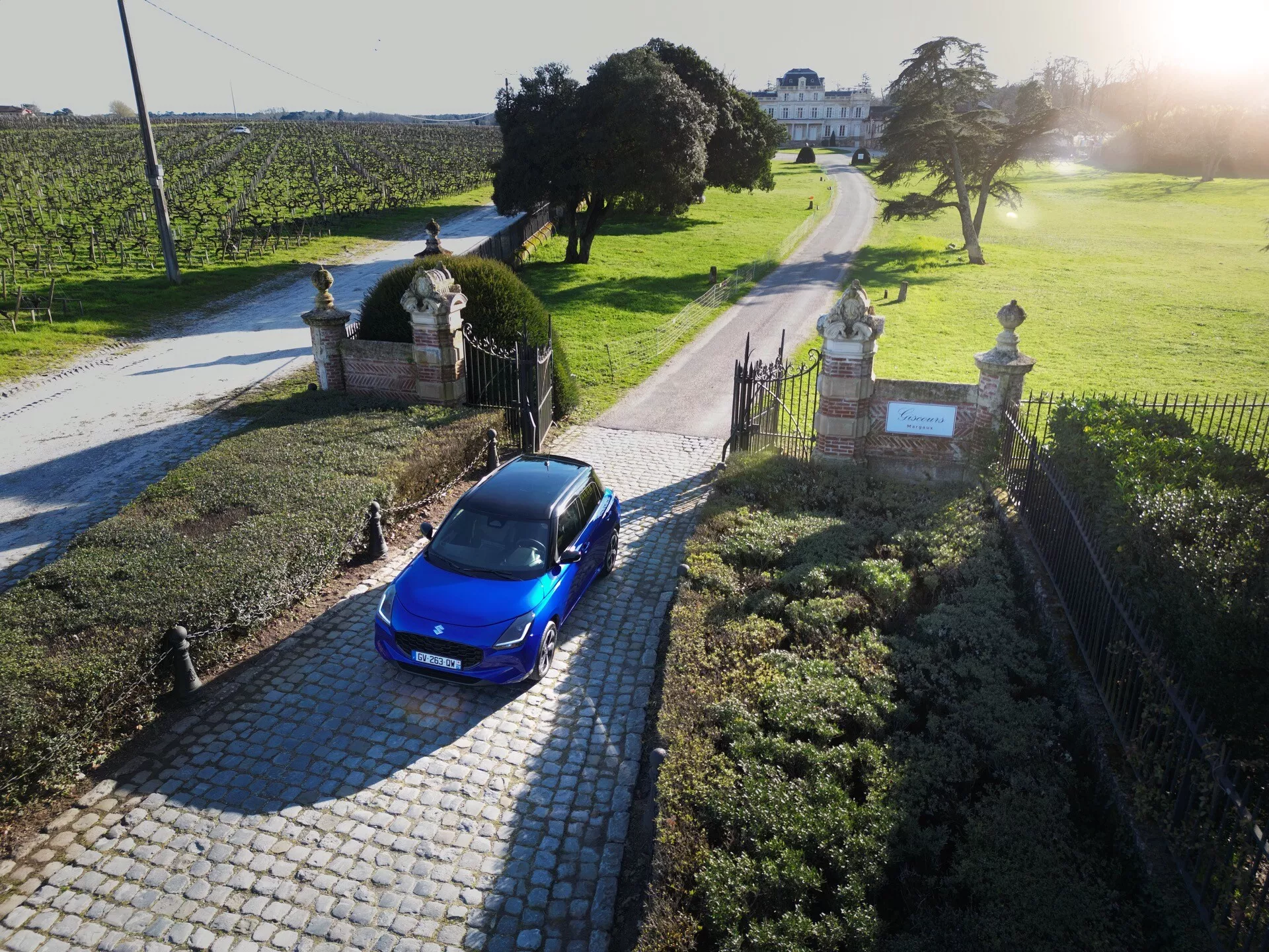 Suzuki Swift 7. generácia 2024 v modrej farbe, pohľad zhora odhaľujúci jej hatchbackový dizajn a LED osvetlenie, zaparkované pri vchode k nehnuteľnosti s upravenou krajinou a vidieckym domom v pozadí.