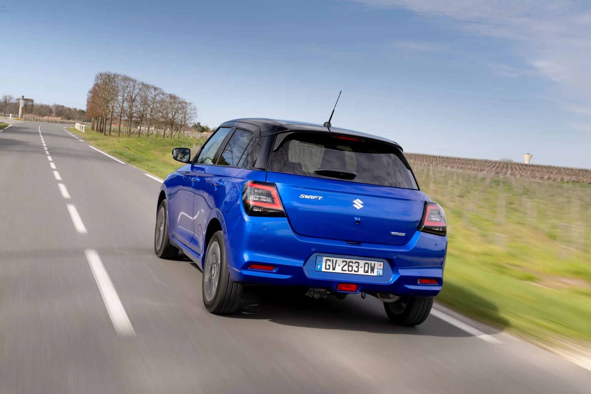 Suzuki Swift 2024 7. generácia v pohybe: Dynamický pohľad zozadu a zo strany na modrý Suzuki Swift odchádzajúci v jasný deň, ukazujúci zadné okno, zadné svetlá, znak Suzuki, dvere na pravej strane, čierny B-stĺpik, spoiler umiestnený na streche, anténu v tvare žraločej plutvy a zliatinové kolesá na vidieckej ceste.