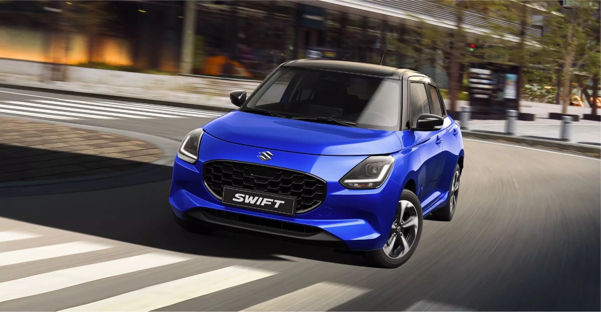 Suzuki Swift 7. generácie 2024 v pohybe, zvýrazňuje ľavú prednú stranu, mriežku, LED svetlomety a dizajn kapoty na pozadí rozostrené mestské ulice