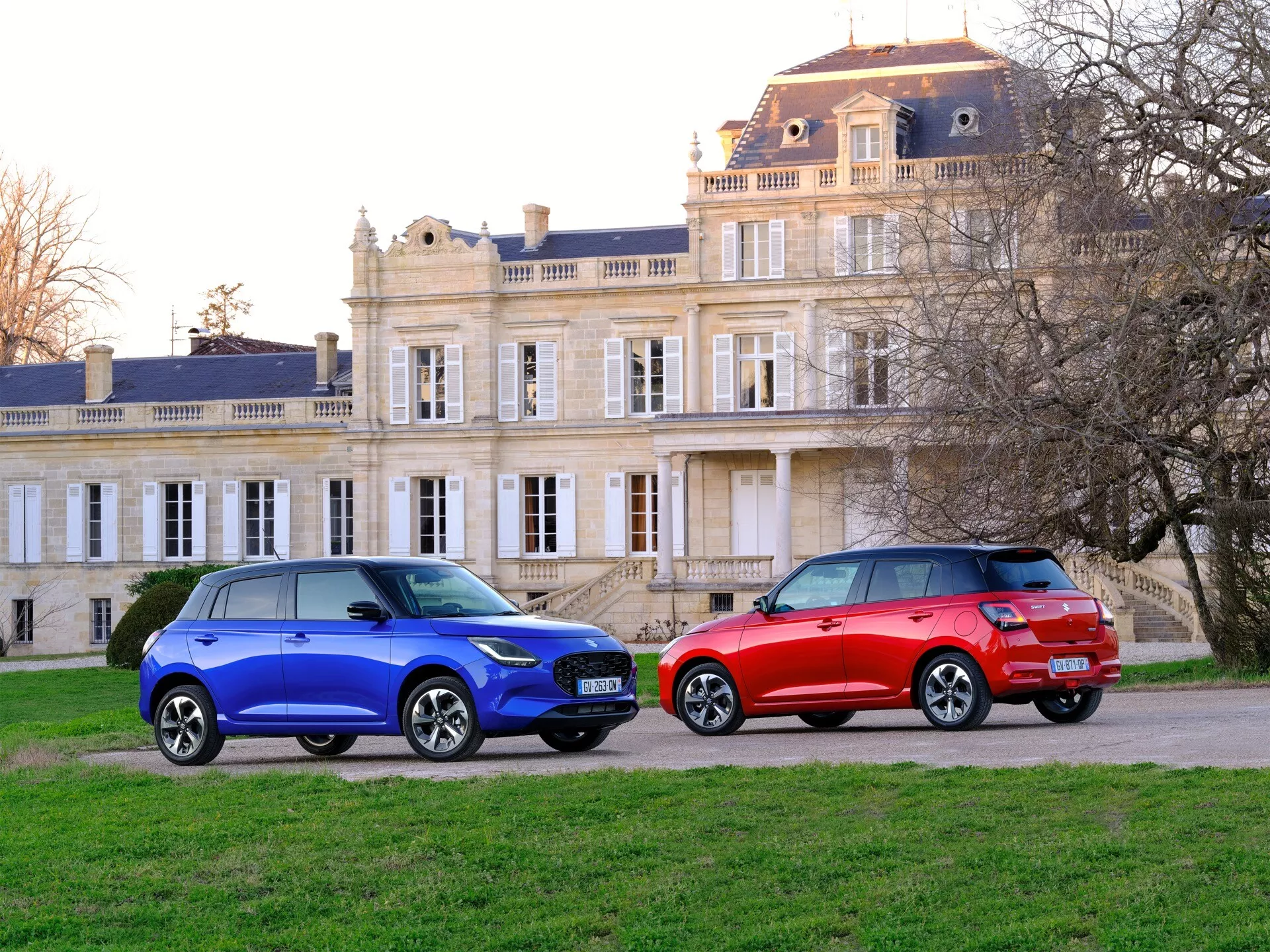 Suzuki Swift 7. generácie 2024 vo vibrantnej modrej a červenej, zaparkované vedľa seba na štrku s historickou budovou v pozadí, ukazujúce nízky uhlový trojštvrťový pohľad na zdôraznenie ich crossover SUV karosérie, zliatinových kolies a moderných predných a zadných svetiel proti pokojnému, európsky inšpirovanému prostrediu.