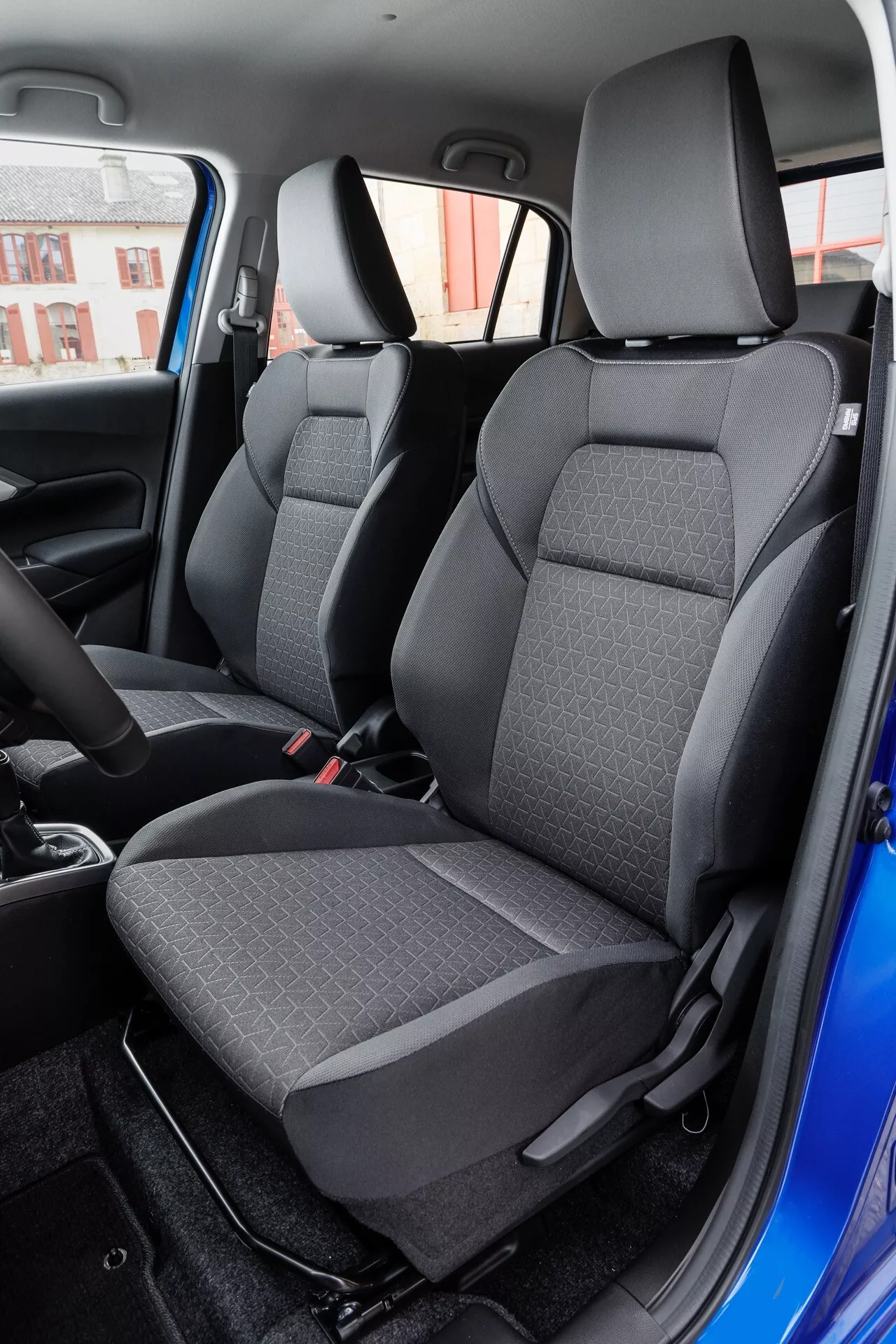 Interiér Suzuki Swift 2024 s pohľadom na predné sedadlá z dvojfarebnej látky z perspektívy zadného pasažiera, zdôraznenie konfigurácie ľavej strany a mestského pozadia