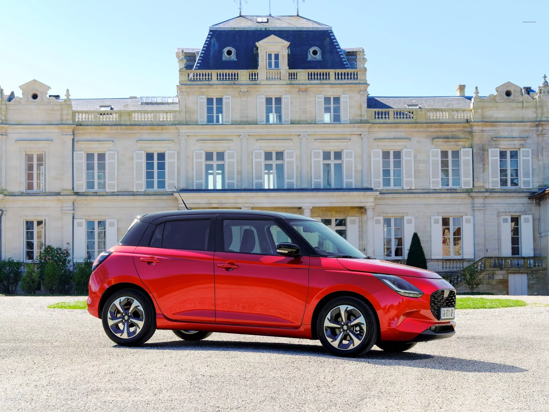 Suzuki Swift 7. generácia ročník 2024 v svetlo červenej farbe, zobrazuje bočný a predný profil s dynamickým aerodynamickým dizajnom a čiernou kontrastnou strechou, zaparkované pred veľkolepou historickou budovou