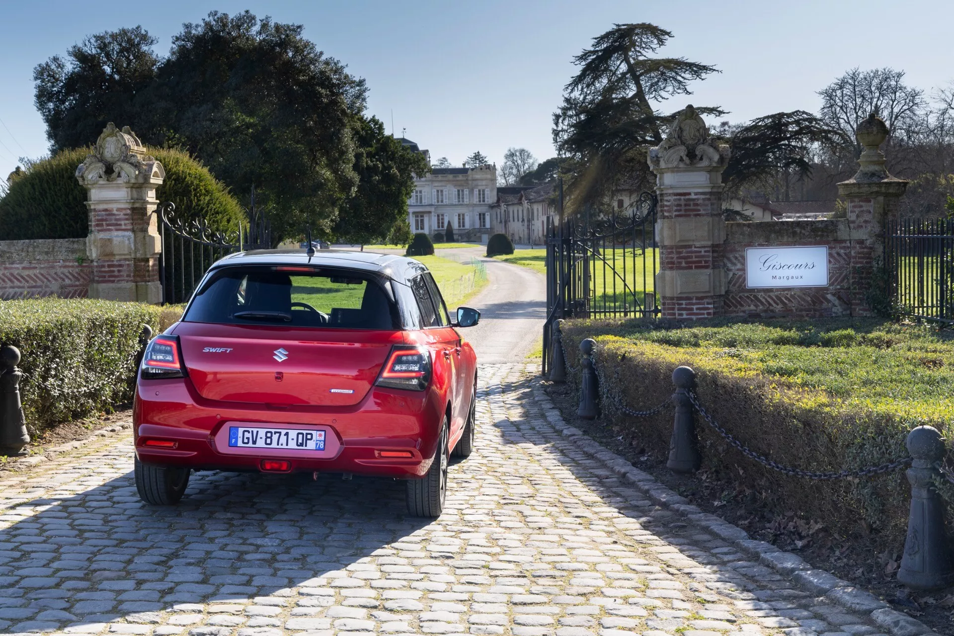 Suzuki Swift 7. generácie ročník 2024 v červenej farbe, zobrazujúci zadné dizajnové prvky vrátane zadných svetiel, zadného okna so stieračom, vysoko umiestneného brzdového svetla, centrálny emblém značky a názov modelu, registračnú tabuľku, systém parkovacích senzorov na nárazníku a integrovanú výfukovú rúru, zaparkovaný na dlaždenej ceste s ozdobnou bránou a veľkým šľachtickým sídlom na pozadí