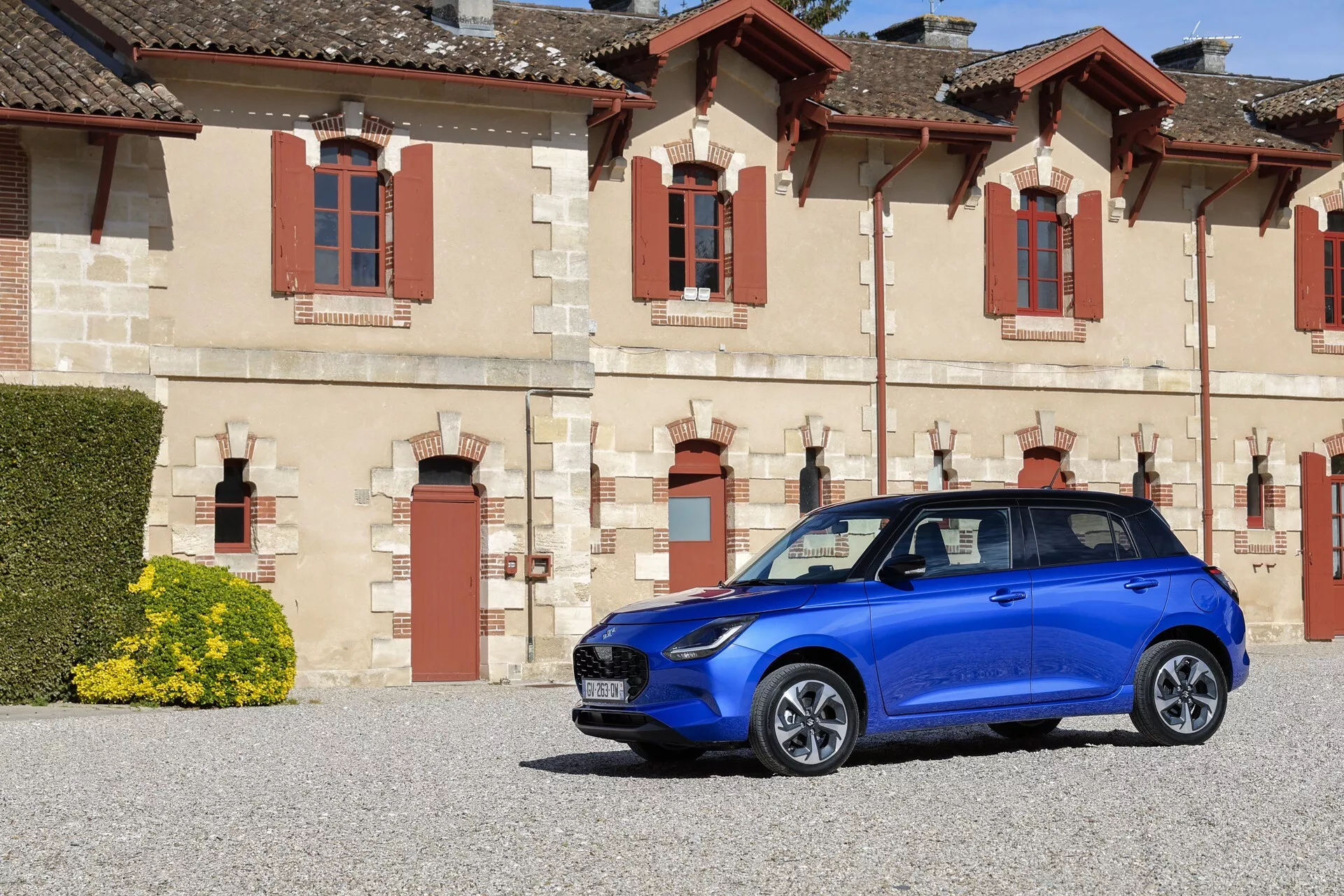 Modré kompaktné auto Suzuki Swift 7. generácie ročník 2024 predstavené v klasickom architektonickom prostredí, zdôrazňujúce prednú mriežku, LED svetlomety, športové kolesá s viacerými paprskami a integrovanú technológiu bočných zrkadiel, zaparkované na štrku s dostatkom priestoru