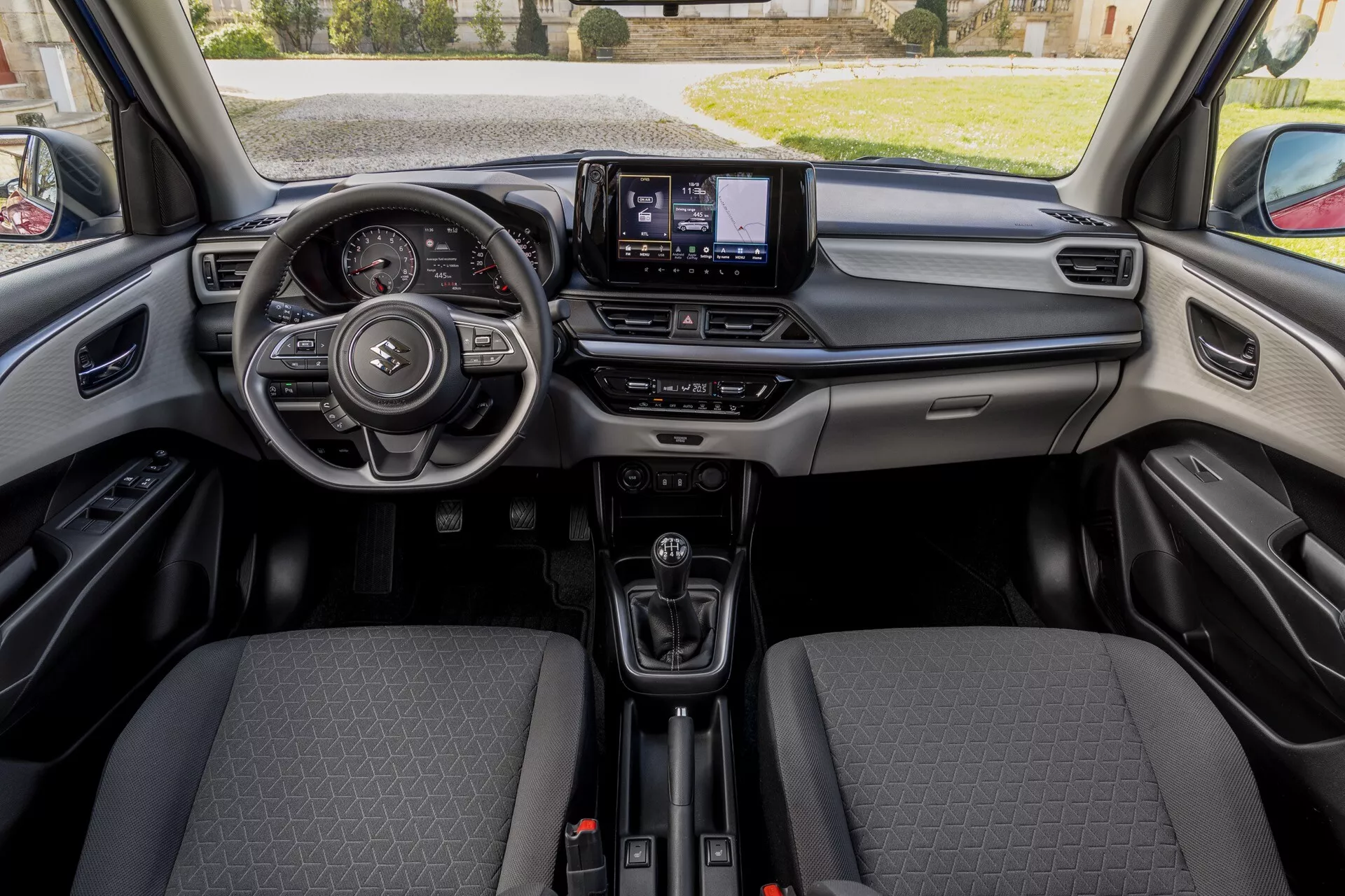 Interiér Suzuki Swift 7. generácie 2024 z pohľadu zo stredu prvej rady, ukazujúci volant s multifunkčnými tlačidlami, elegantnú prístrojovú dosku s potenciálnym digitálnym displejom, centrálny obrazovku infotainmentu, ergonomickú páku radenia, predné sedadlá s látkovým dizajnom a výrazným vzorom, viditeľný interiér dverí s pravdepodobnými ovládacími prvkami, držiaky na poháre a USB/napájacie zásuvky, pod prirodzeným svetlom bez ostrých tieňov