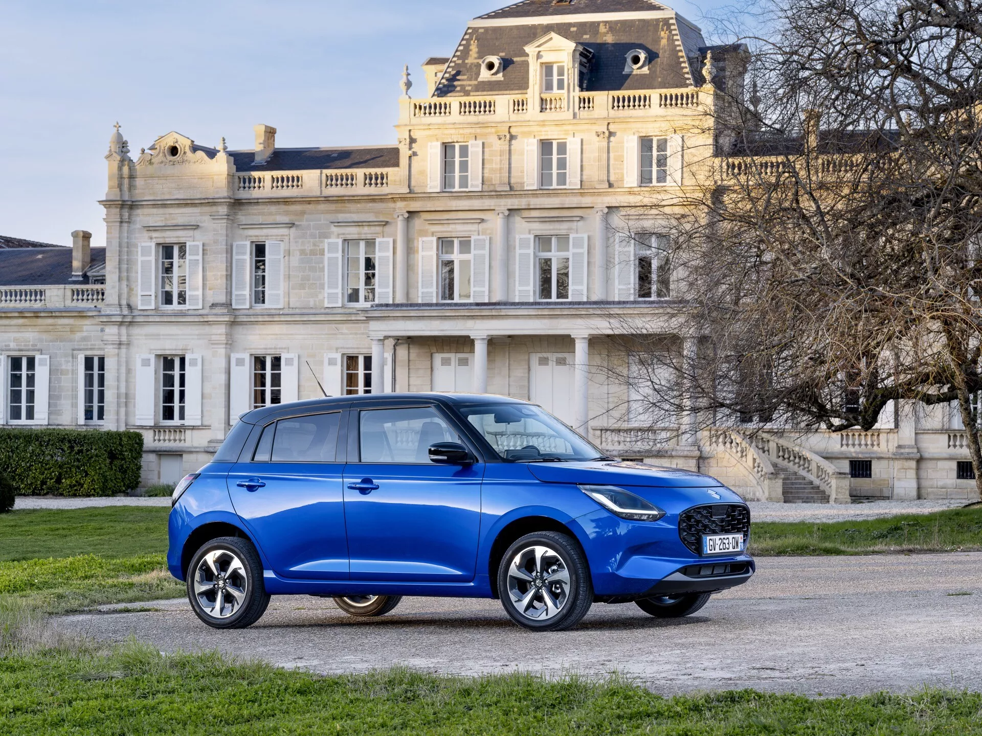 Modré kompaktné SUV Suzuki Swift 2024 zaparkované na príjazdovej ceste s klasickou budovou na pozadí, ukazuje svoj pohľad spredu z troch štvrtín s detailnou prednou mriežkou, LED alebo HID svetlometmi, hmlovými svetlami, kolesami z ľahkej zliatiny s viacerými ramenami a bočnými zrkadlami s integrovanými smerovkami v luxusnom prostredí
