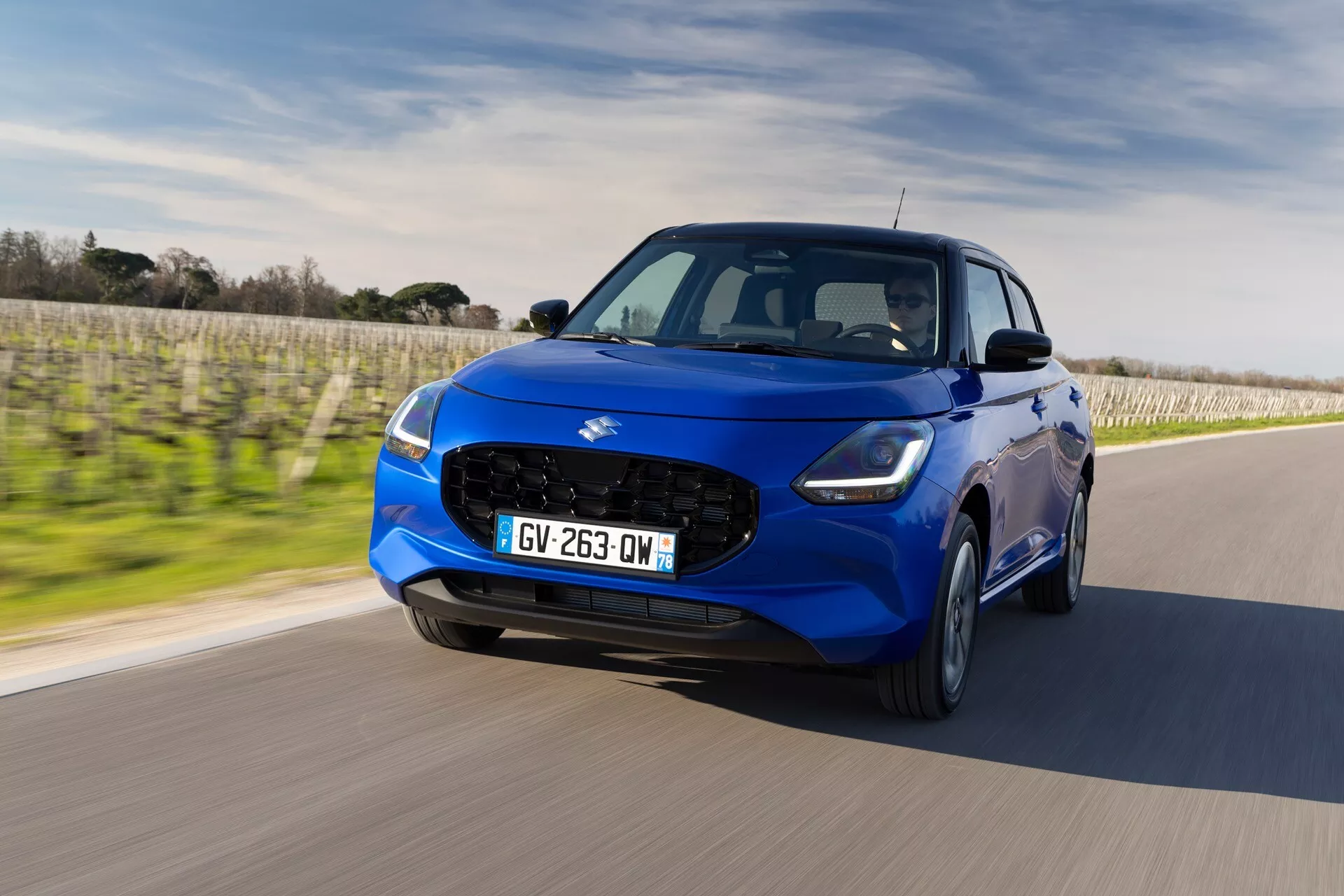 Moderný hatchback Suzuki Swift 7. generácie z roku 2024 v pohybe pod jasným nebom, predvádzajúci elegantný dizajn s výraznou mriežkou, zašpicatými svetlometmi a športovými zliatinovými kolesami