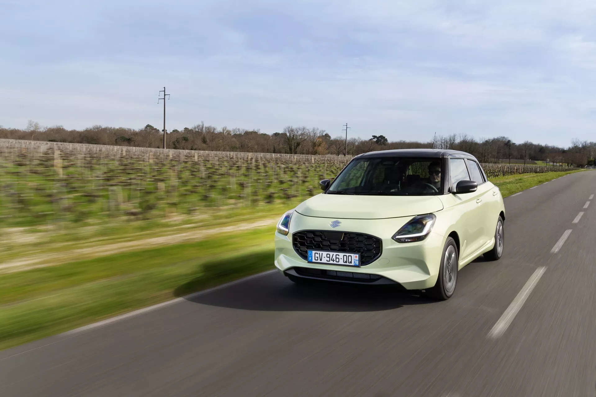 Suzuki Swift 7. generácie 2024 v dynamickom pohybe, zobrazenie z uhla trištvrte prednej časti ukazuje dizajn elektrického kompaktného auta, modernú prednú fasádu a bočný profil proti rozmazanému pozadiu, zdôrazňuje rýchlosť a modernosť
