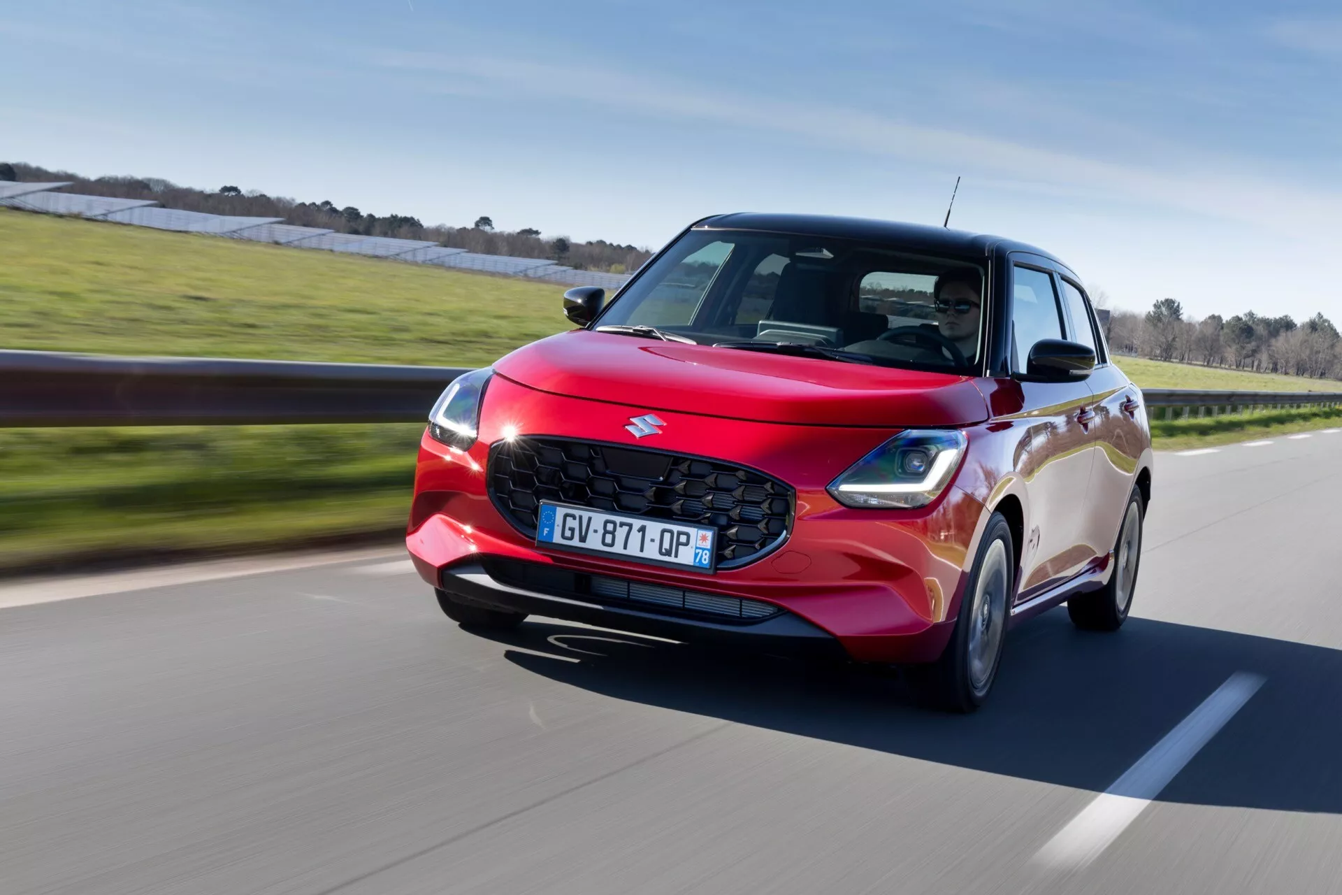 Suzuki Swift 2024, 7. generácia vo svetlo červenej farbe, predvádza lesklý povrch a moderné vlastnosti vrátane LED svetlometov a zliatinových kolies, na dobre udržiavanej ceste obklopenej zeleňou, zachytené z dynamického uhla tri štvrtiny spredu