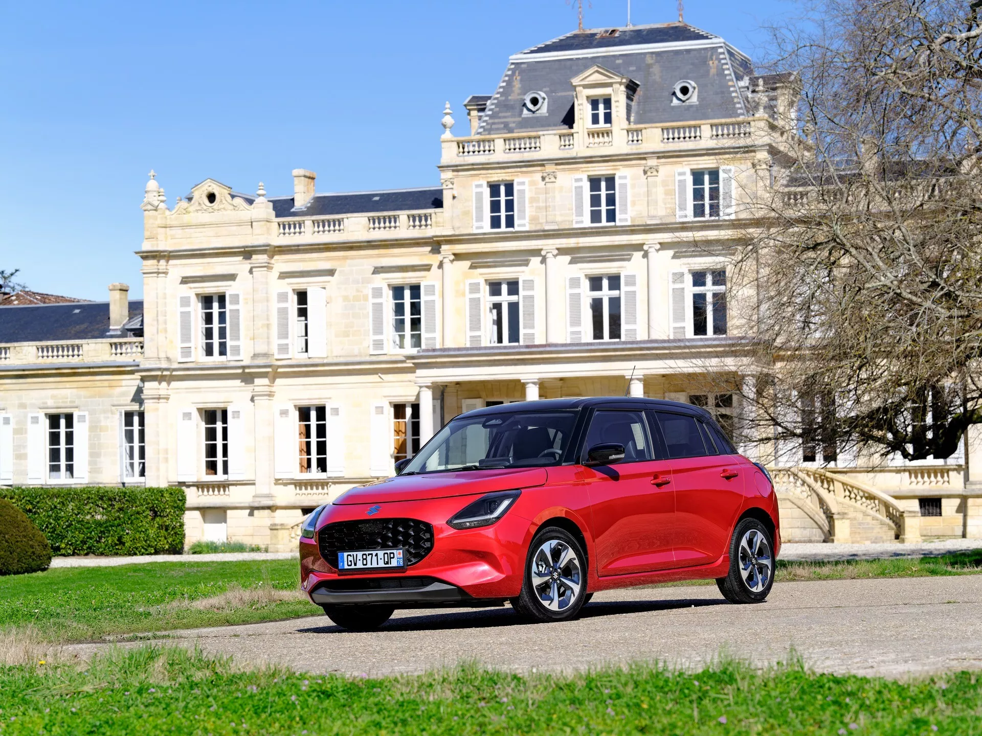 Suzuki Swift 2024, 7. generácia, v jasne červenej farbe zaparkované vonku pred klasickou budovou, zobrazujúce jeho pohľad z prednej strany zo štyroch tretín so zreteľným dôrazom na prednú mriežku, LED svetlomety, dizajn predného a zadného kolesa a športové nízkoprofilové pneumatiky proti pozadiu bohatého prostredia, zdôrazňujúce moderné dizajnové prvky a možno pokročilé funkcie ako hybridný alebo elektrický pohon.