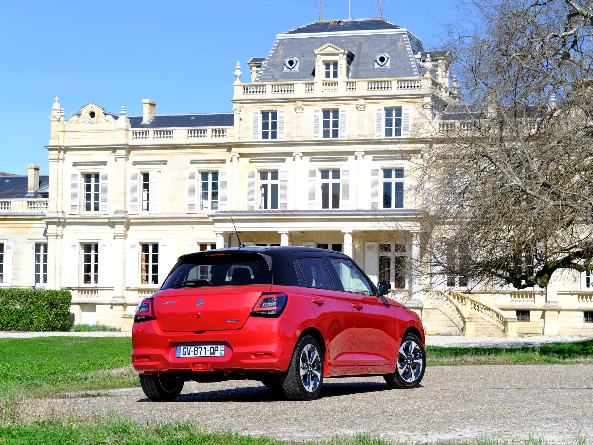 Červený hatchback Suzuki Swift 2024 s moderným dizajnom z trojštvrťovej zadnej perspektívy ukazujúci bočné a zadné profily, proti elegantnému klasicistnému sídlu s upraveným areálom, zdôrazňujúce luxusný životný štýl a pokročilú automobilovú technológiu.
