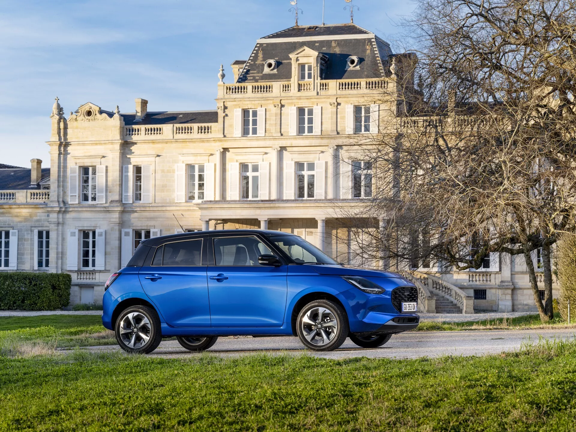 Suzuki Swift 7. generácie 2024 v elektricky modrej farbe s aerodynamickým dizajnom, LED svetlomety, výraznou mriežkou, viacramennými zliatinovými kolesami a integrovanými smerovkami na bočných zrkadlách na pozadí s trávnatým luxusom
